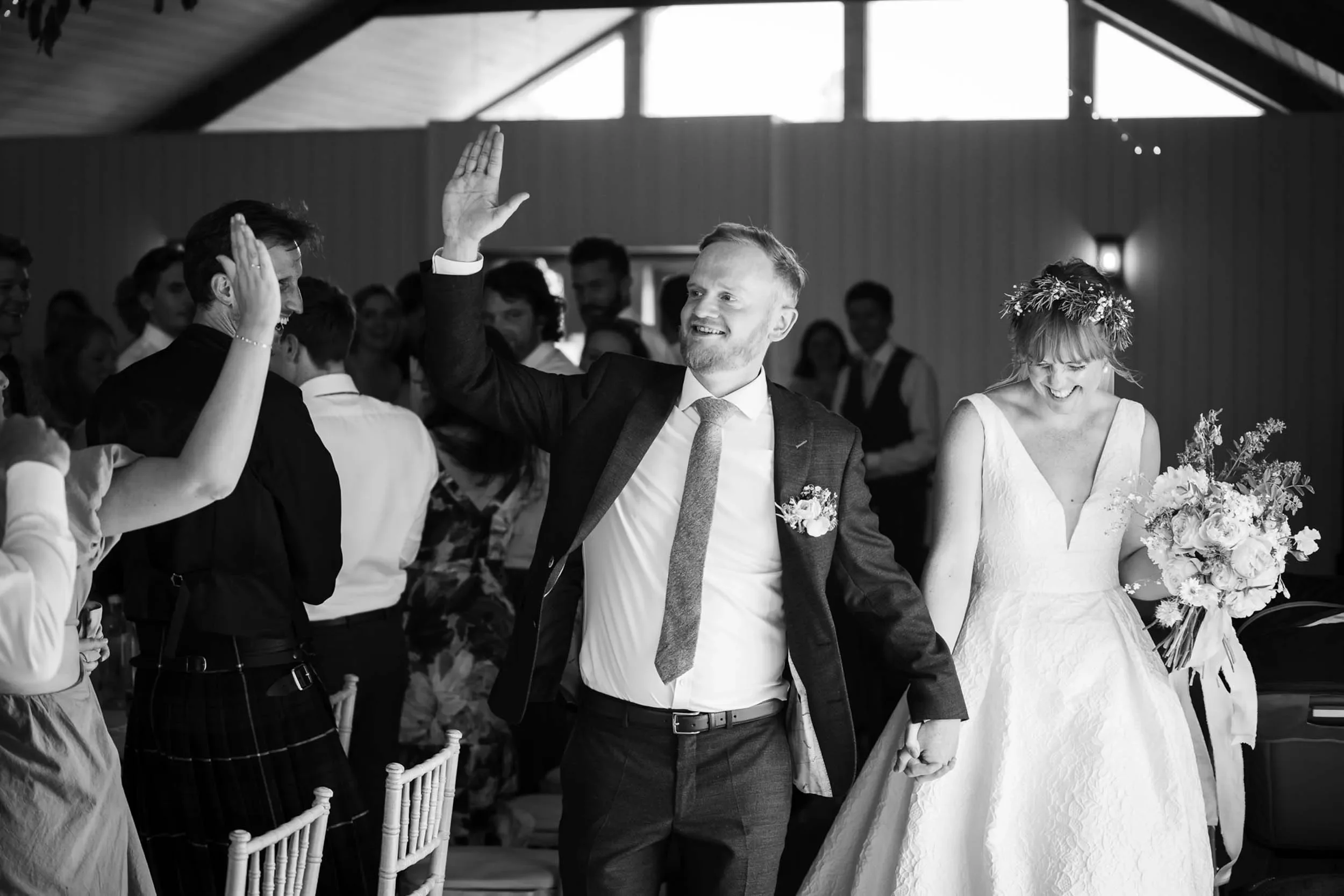 A groom in a suit with a tie and boutonniere high-fives a guest at his wedding reception. The bride in a white wedding dress with a floral crown and bouquet stands nearby, smiling. Guests are seen in the background inside a venue with wooden walls an