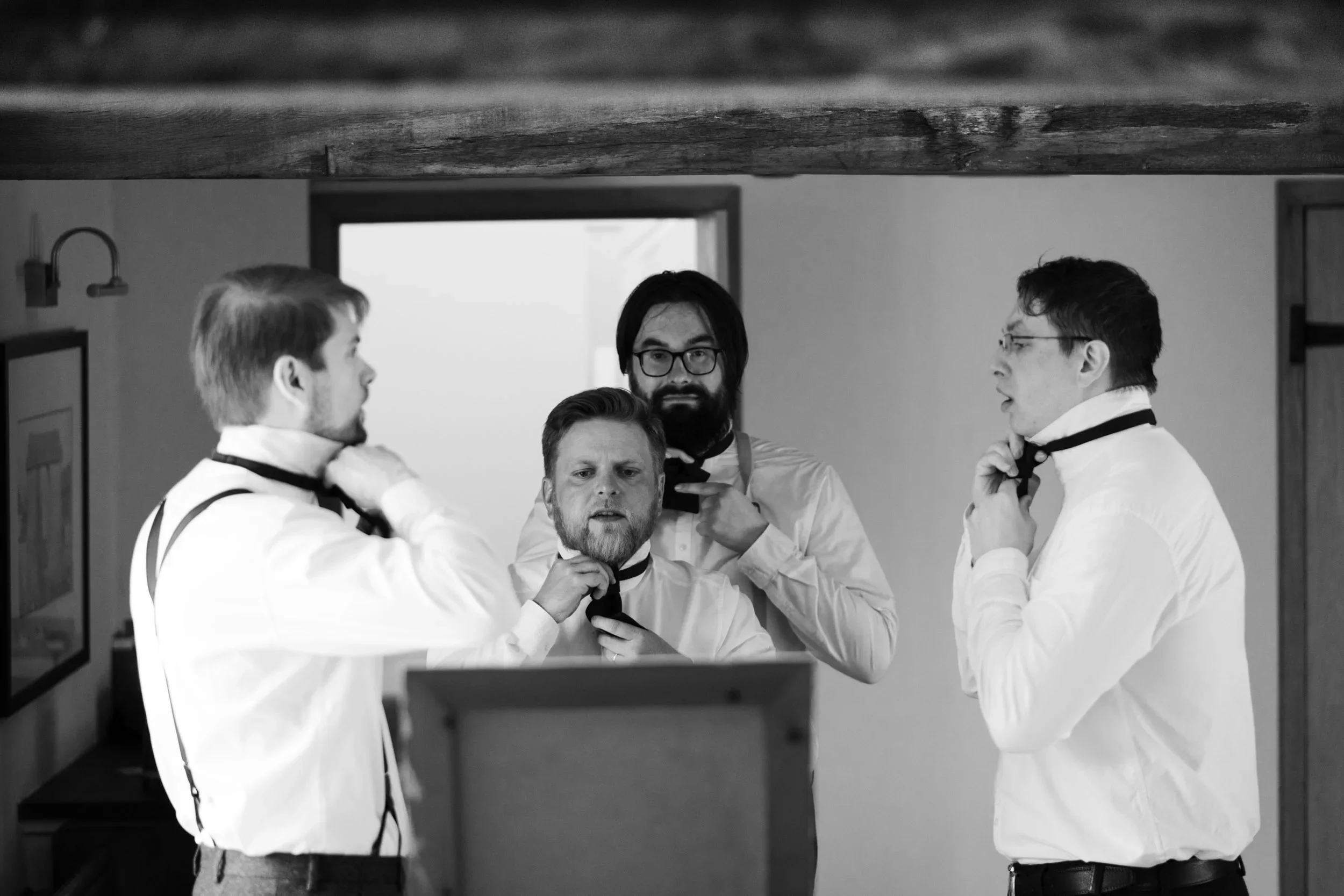Four men dressed in formal white shirts and black bow ties adjusting their bow ties in front of a mirror in a room with wooden support beams.