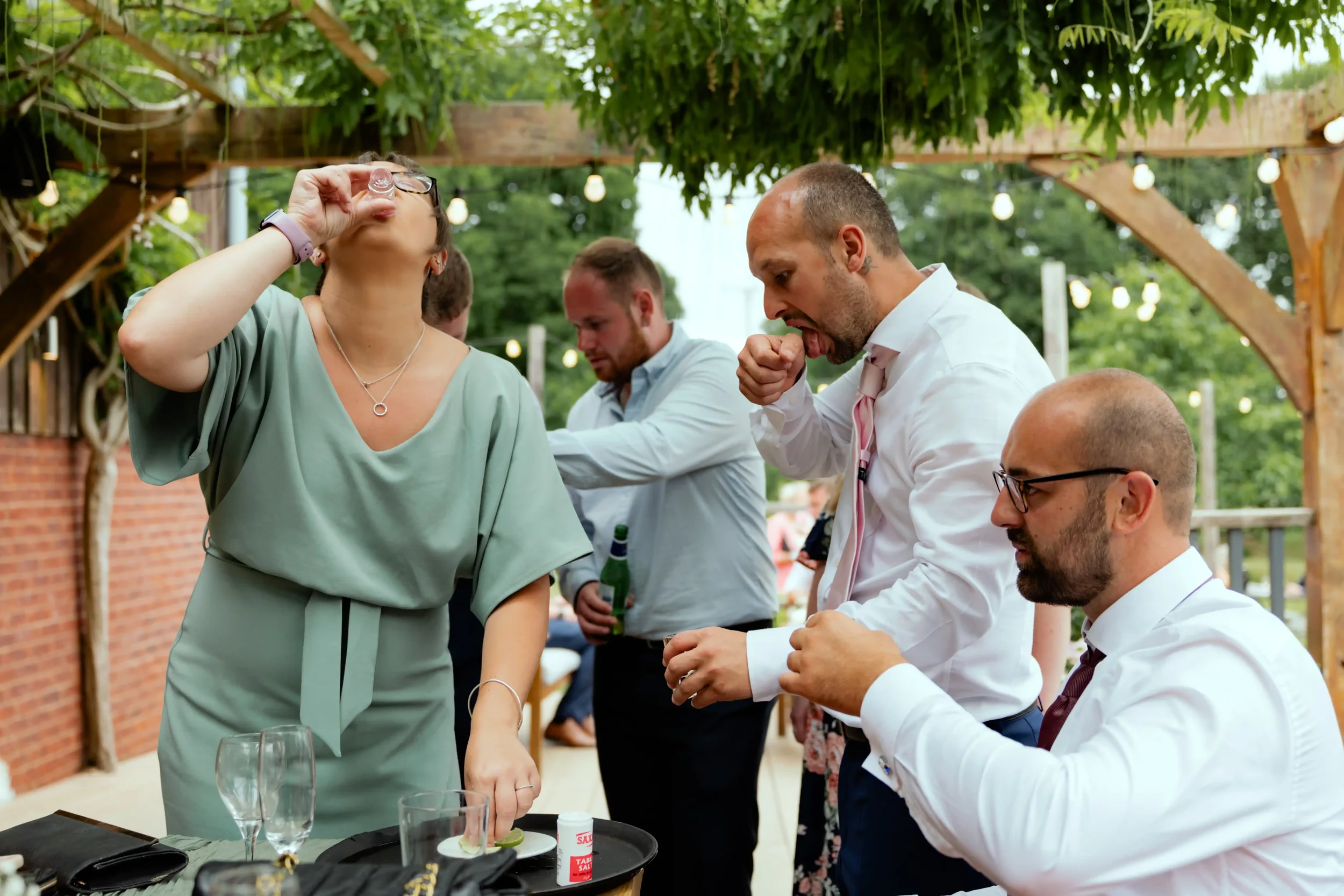 People at an outdoor party, some seated and some standing, drinking and socializing under a wooden pergola with string lights. Women and men are dressed in semi-formal attire, with glasses, bottles, and a small tray of drinks visible.