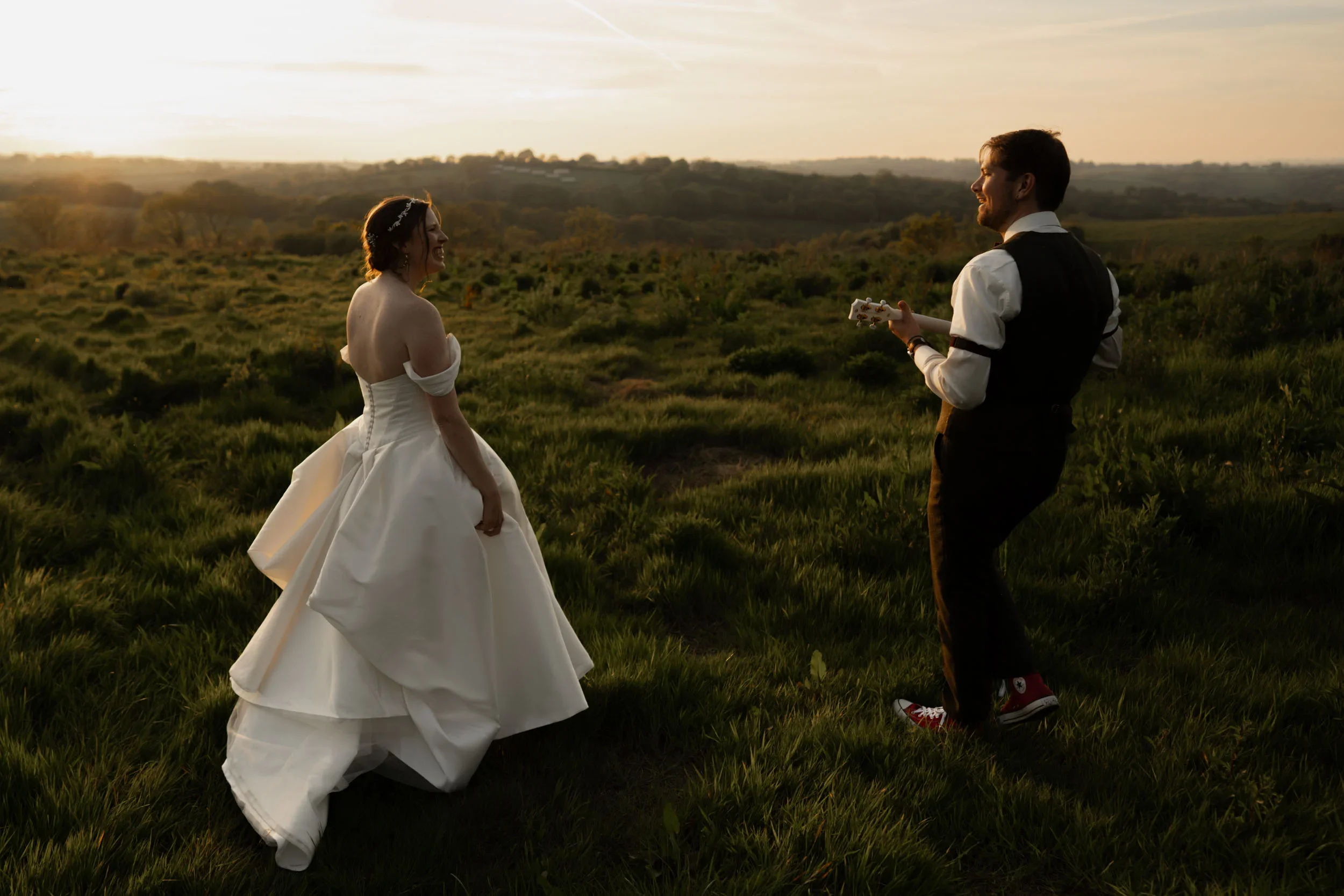 A bride in a white wedding gown and a groom in a suit with red sneakers standing in an open field at sunset, facing each other with the groom playing a guitar.