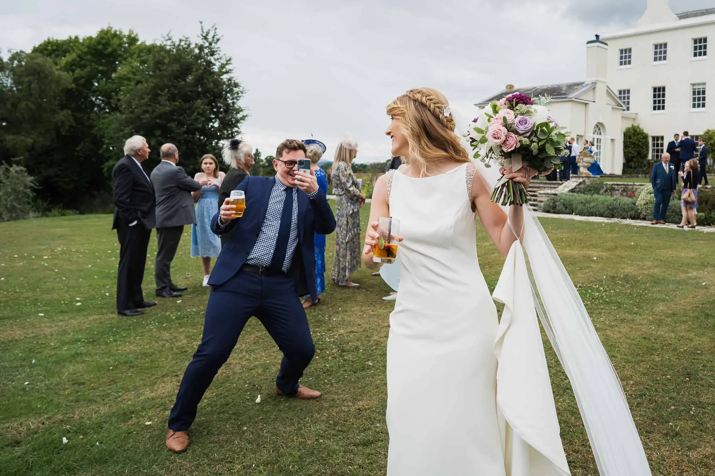 A woman in a white wedding dress holding a bouquet of pink and purple flowers stands with a drink in hand, smiling at a man in a dark suit and glasses, who is taking a picture of her with his phone. Several other guests are in the background on a gra