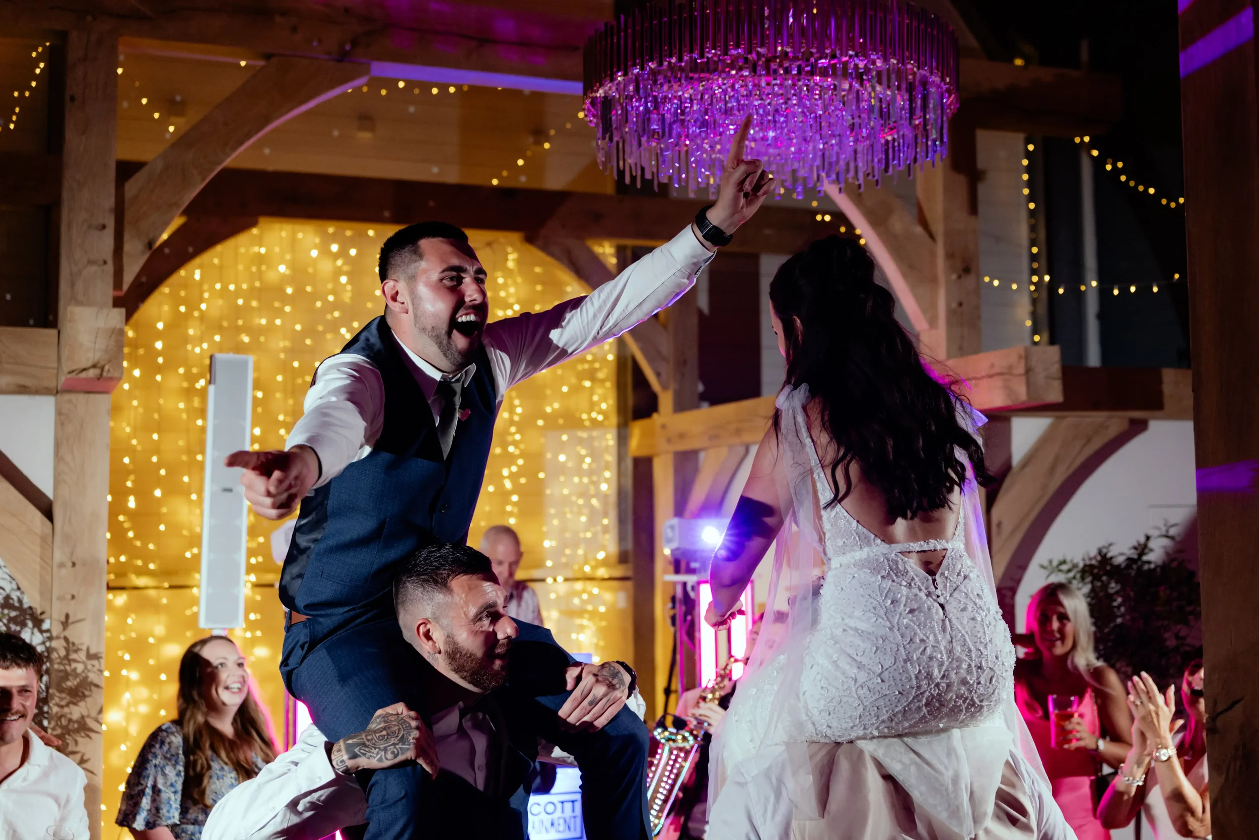 People celebrating at a wedding reception, with one man standing on another man's shoulders pointing his finger, and a woman in a white dress facing them, in a decorated venue with warm lighting and string lights.