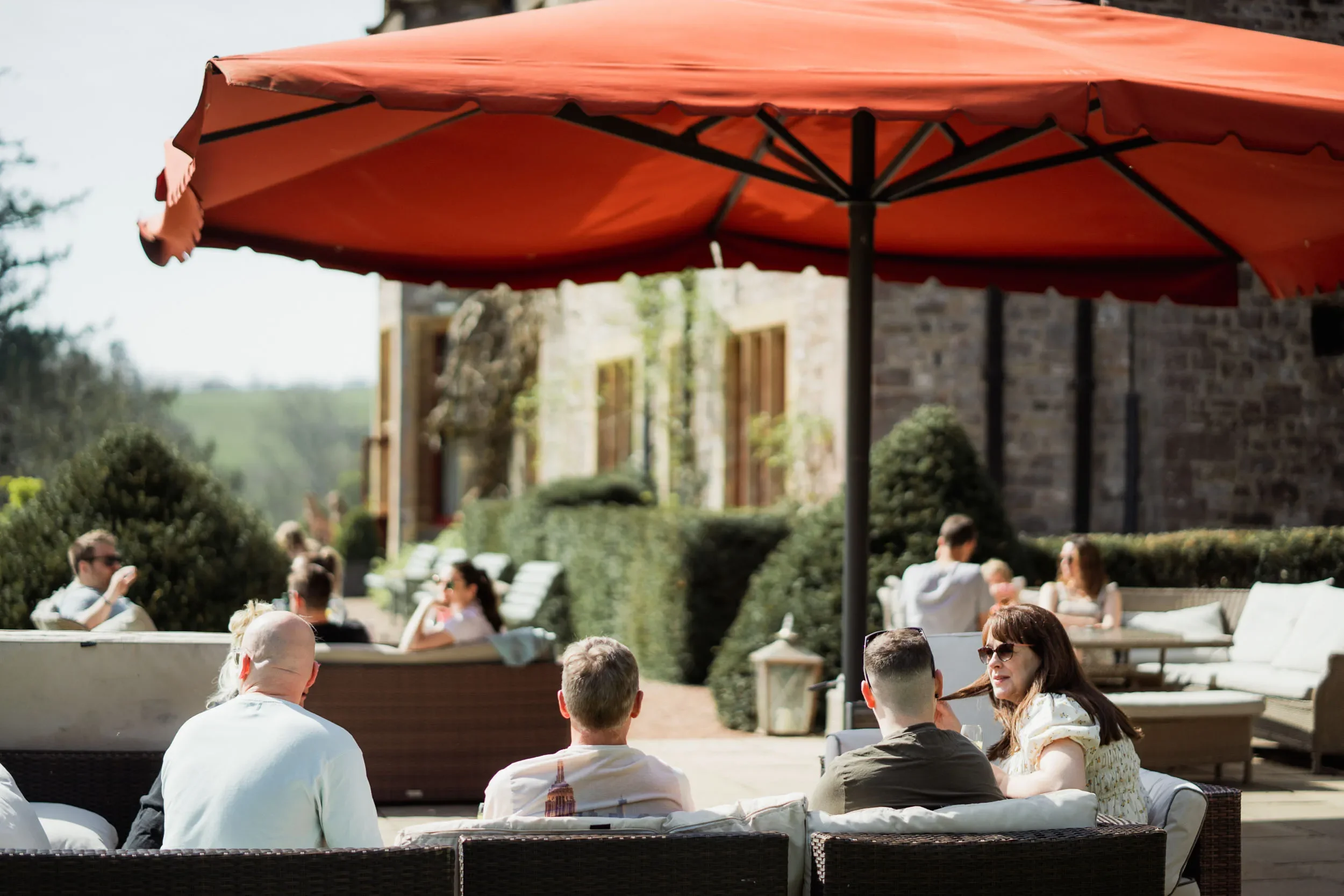 People sitting and socializing outdoors under a large red patio umbrella at a garden party or outdoor cafe.