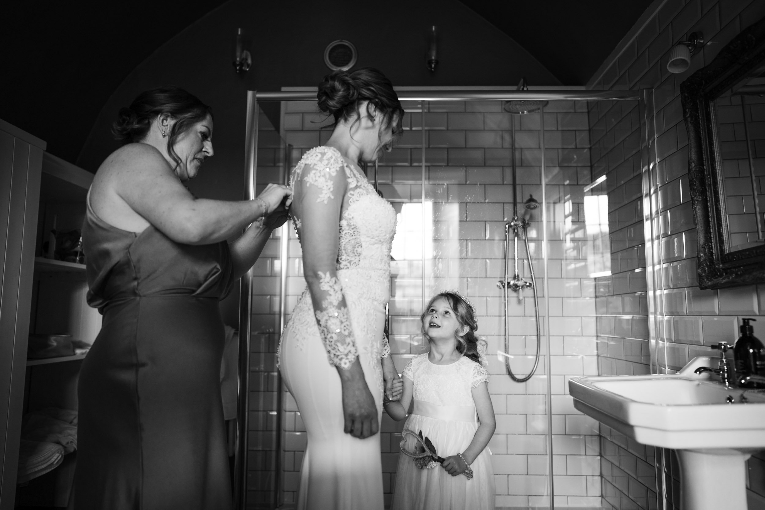 A bride in a lace wedding dress is being helped into her gown by a woman while a young girl in a white dress looks up at her with a smile in a tiled bathroom.