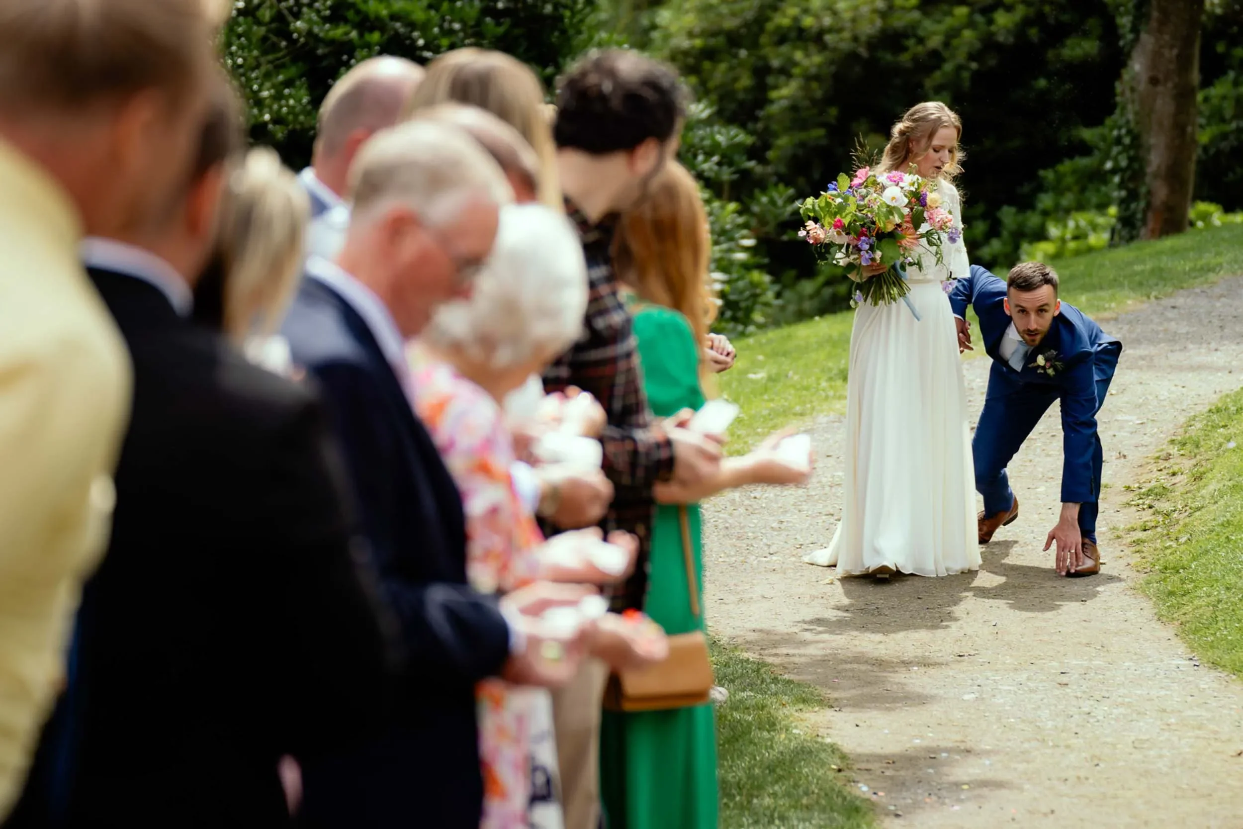 A wedding ceremony outdoors with a line of guests, a bride holding a bouquet, and a groom bending down on a dirt path surrounded by greenery.