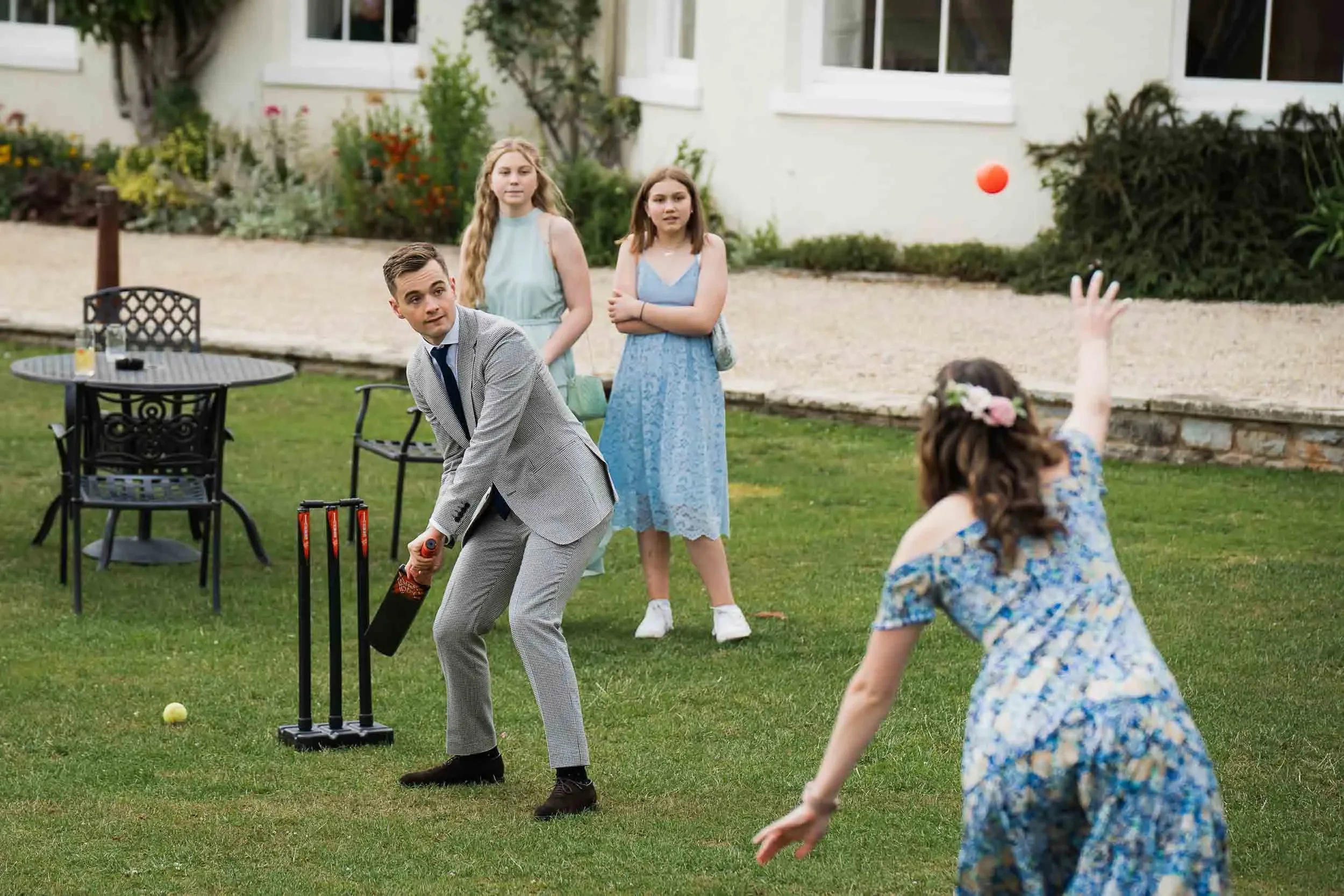 A man in a gray suit preparing to bowl, with four young girls watching as one girl throws a red ball in a yard with green grass and outdoor furniture.