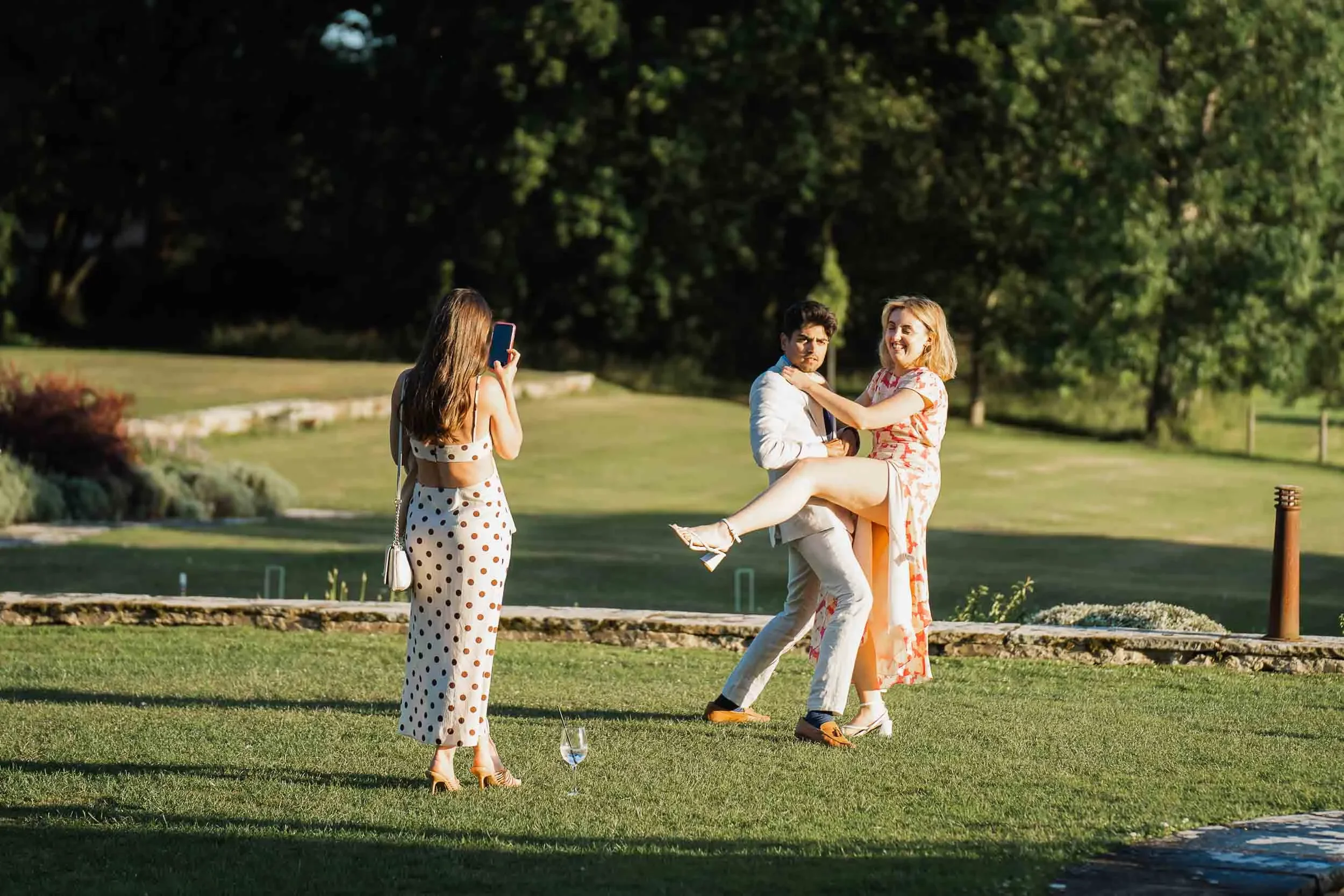 A man and woman, dressed in formal clothes, are dancing outdoors while another woman takes a photo of them with her phone. The scene is set in a green park or garden with trees and grass.