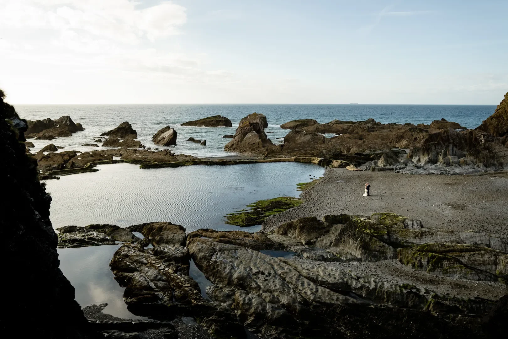 A couple in wedding attire standing on a rocky beach near a tidal pool with the ocean in the background.
