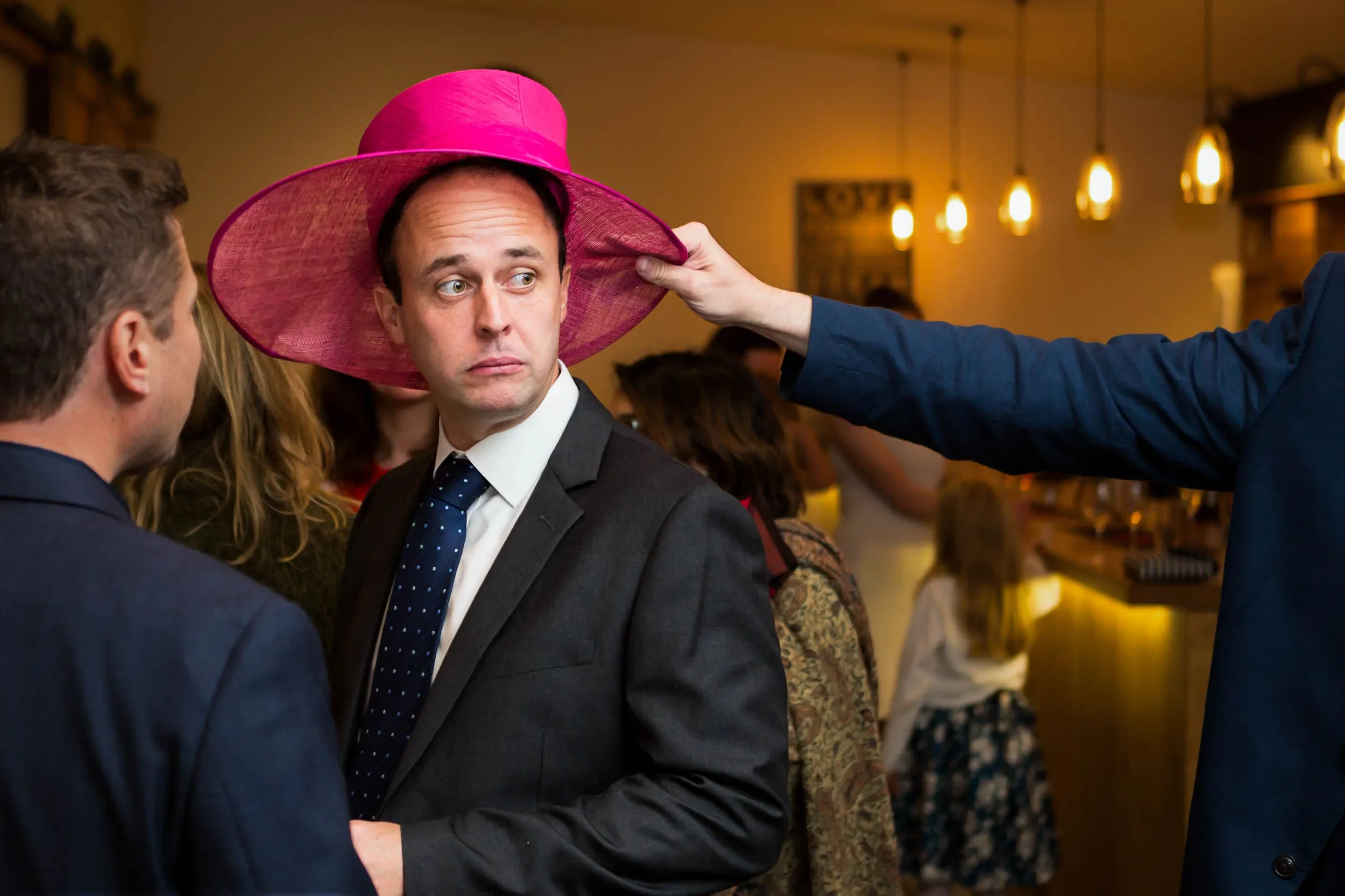 A man in a suit and tie wearing a large pink hat being adjusted by another person at a social gathering in a warmly lit indoor space.