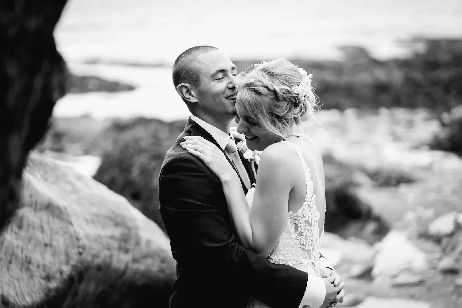 Black and white photo of a smiling couple, dressed in wedding attire, embracing outdoors near water and rocks.