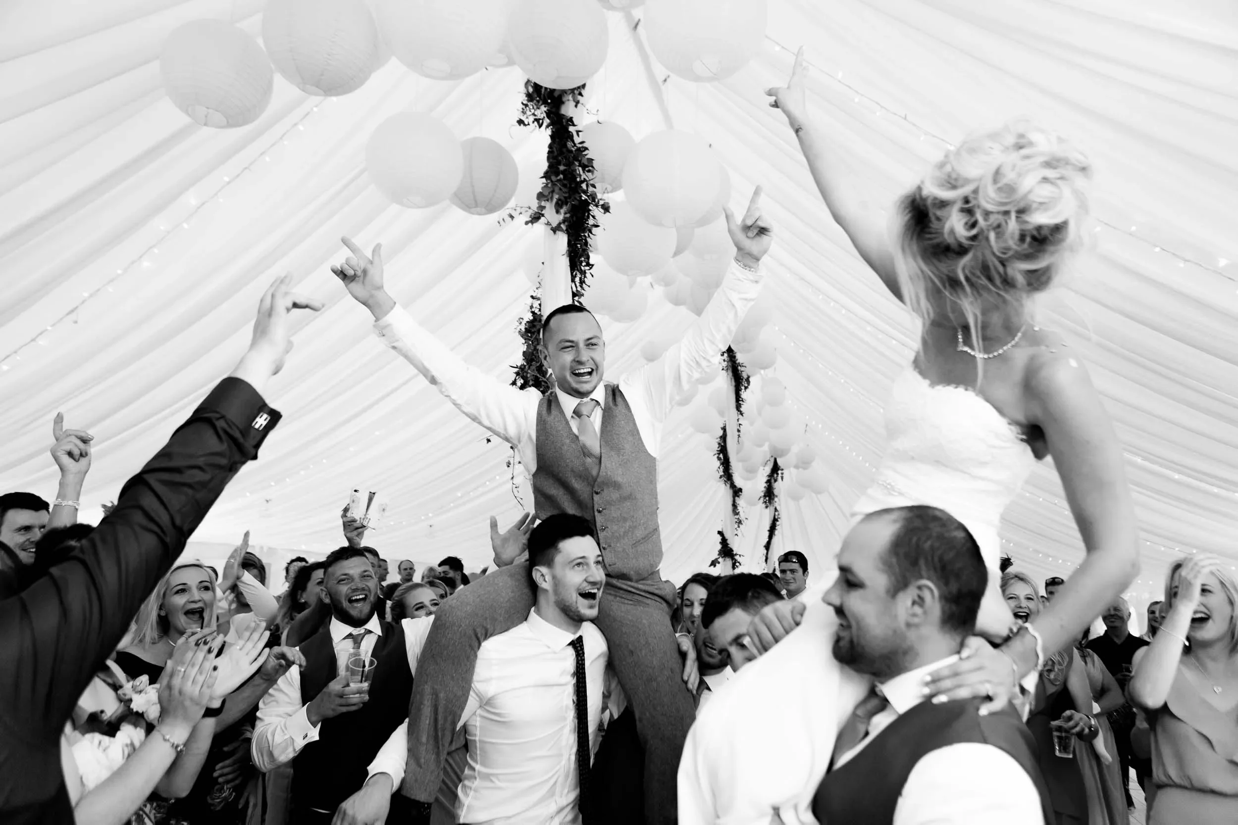 A lively wedding celebration with a bride and groom surrounded by guests, as the groom is carried on the shoulders of another guest inside a decorated tent with hanging paper lanterns.
