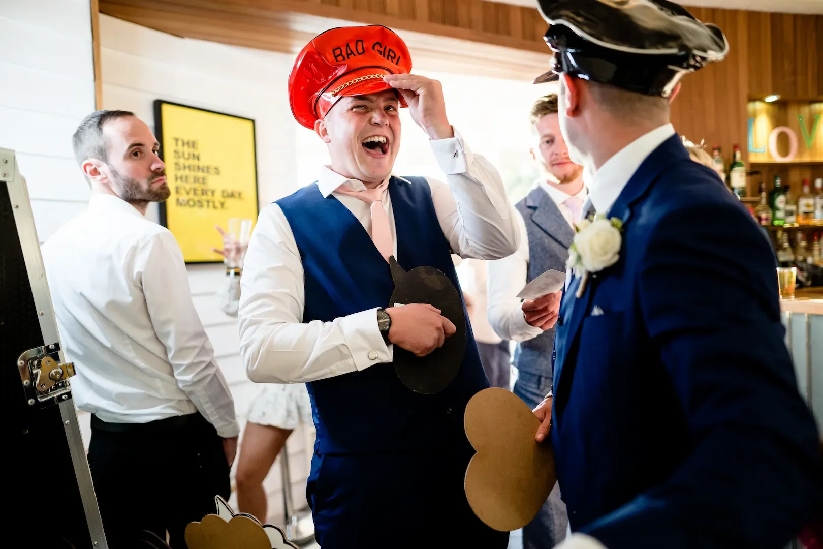 Four men at a party, one wearing a red hat labeled 'Bad Girl,' and another in a blue suit and police cap, engaged in conversation, with a bar in the background.