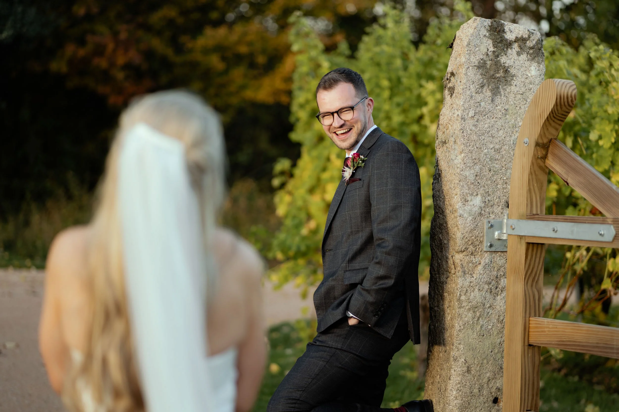 A groom in a dark suit with glasses and a boutonnière, smiling and leaning against a stone pillar outdoors during a wedding, with a blonde bride out of focus in the foreground.