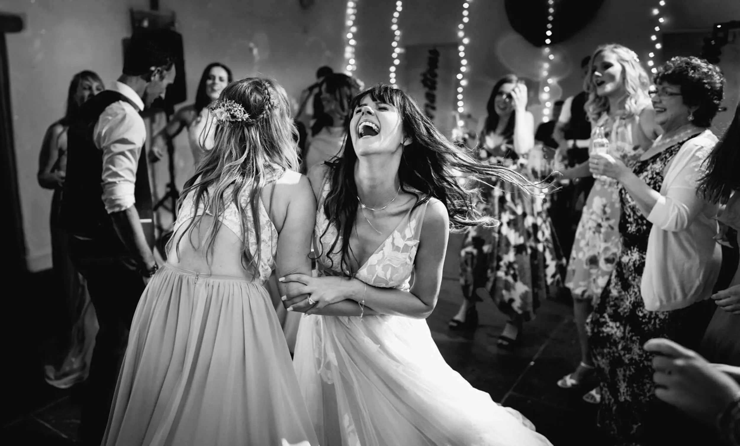 A group of women dancing and celebrating at a party, with some holding drinks and smiling, illuminated by string lights overhead, in a lively social setting.
