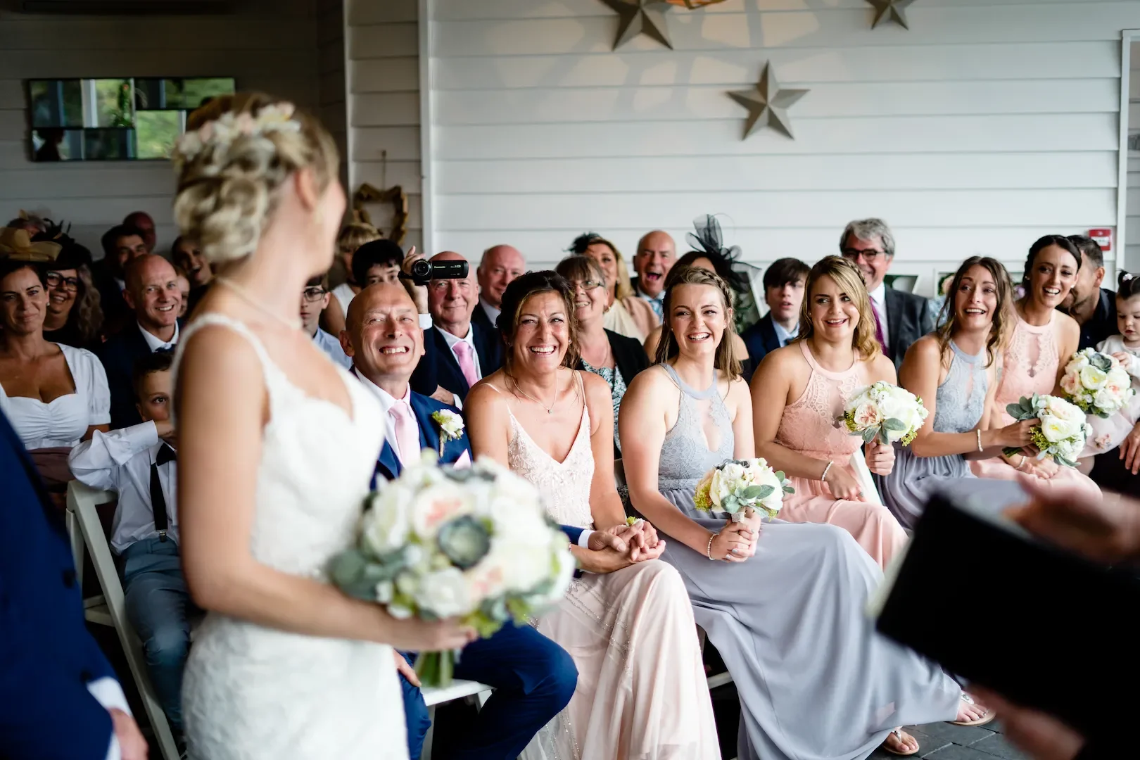 Bride holding a bouquet at her wedding ceremony with guests smiling and laughing in the background.