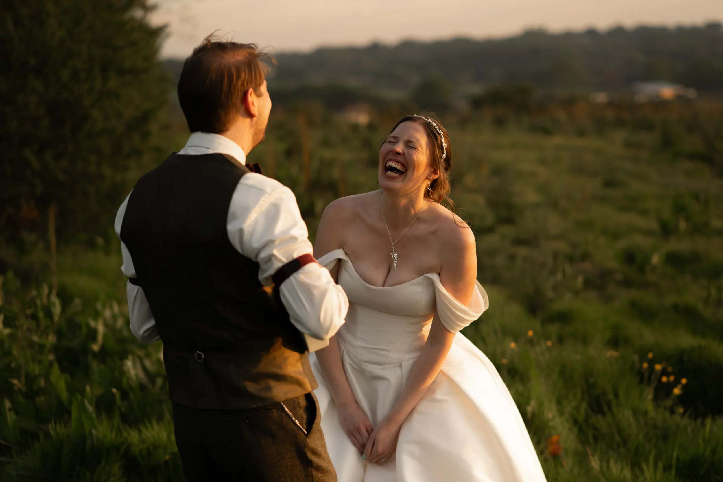 A woman in a wedding dress laughing joyfully with a man in formal attire on a scenic outdoor setting during sunset.