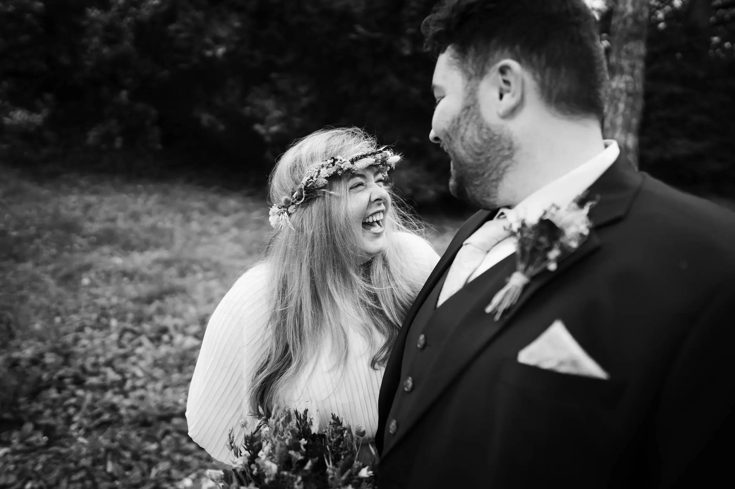 A woman with long hair, wearing a floral crown and a light-colored dress, laughing joyfully while looking at a man in a suit with a boutonniere, who is smiling at her. They are outdoors in a natural setting.
