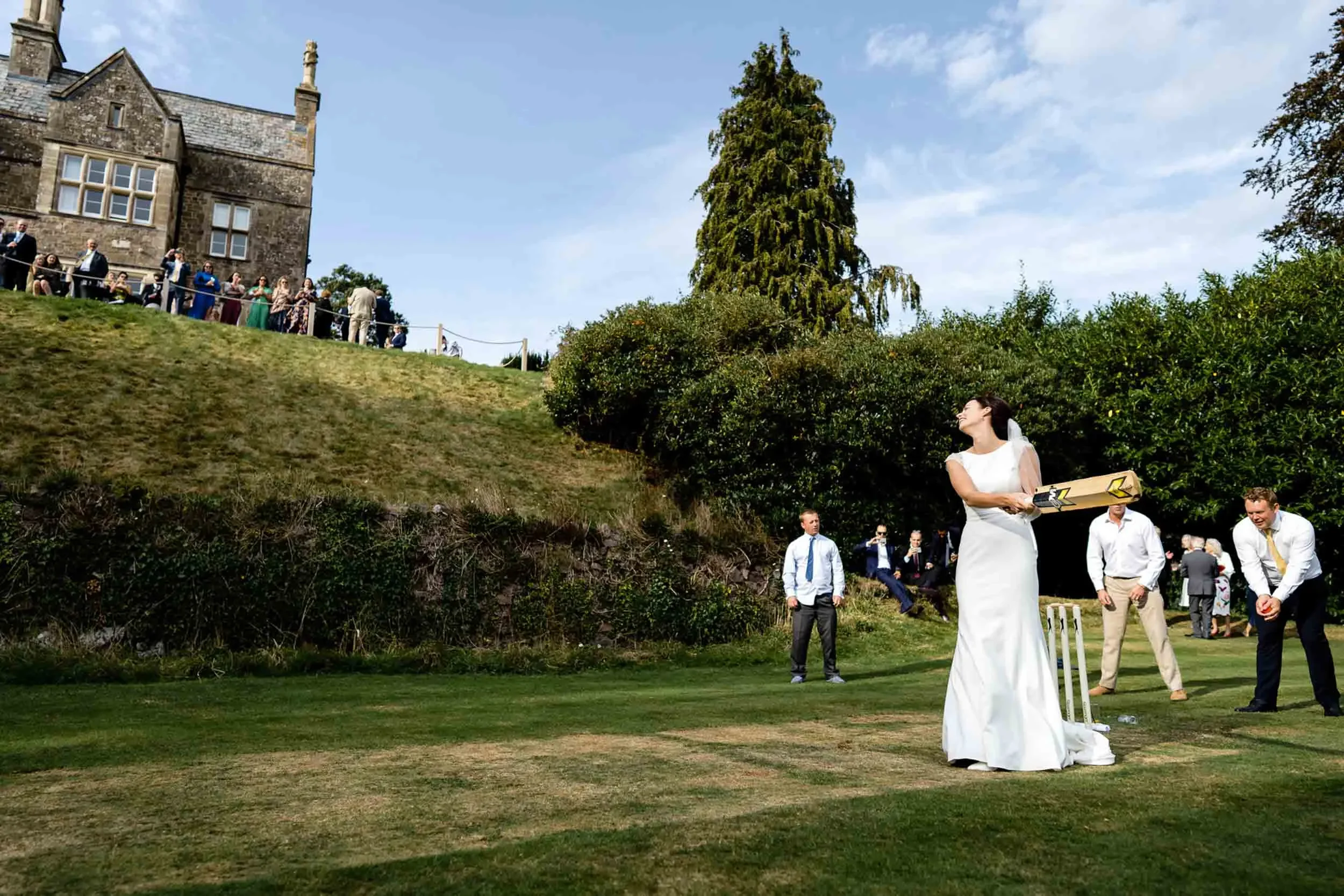 Wedding reception on a lawn with a bride in a white dress holding a cricket bat and a man in a white shirt and tie preparing to bowl. Guests are sitting and standing on a hill, watching the scene. A large house is in the background, surrounded by gre