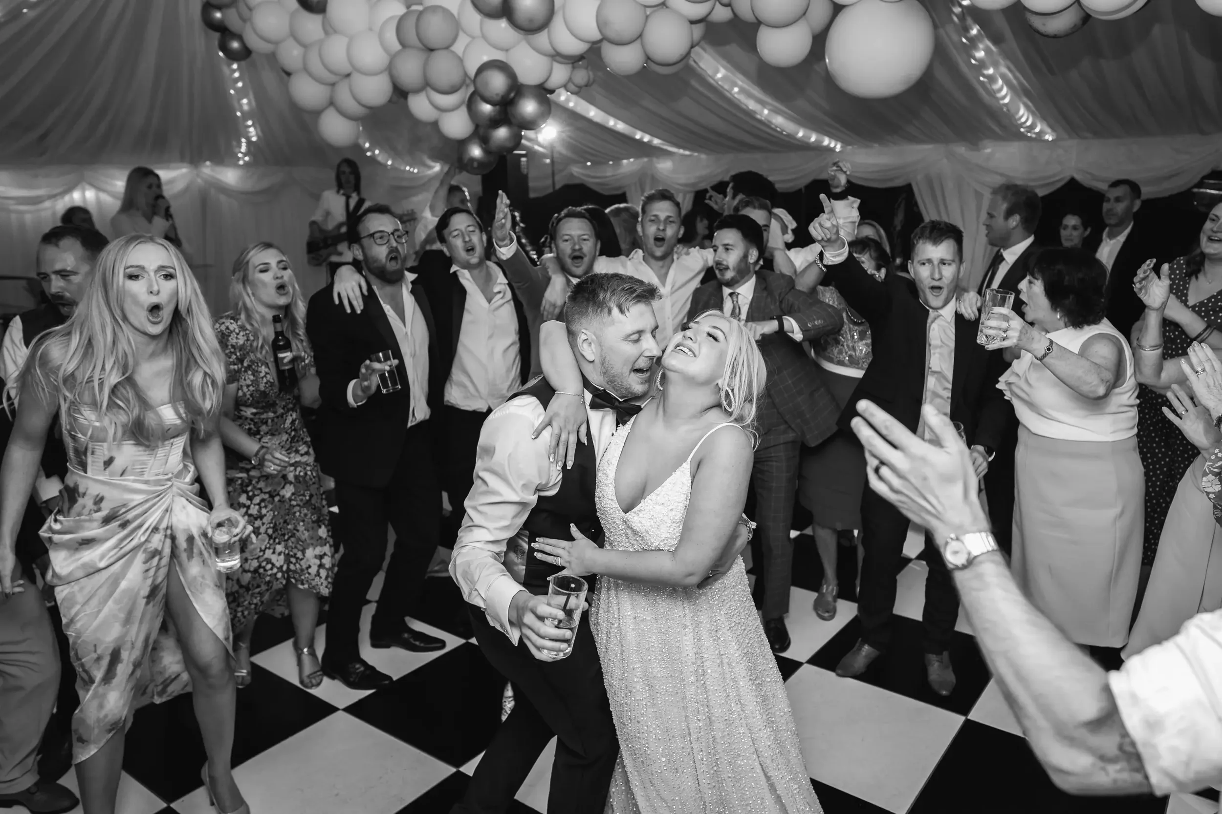 A black and white photo of a lively wedding reception dance floor. A couple in the center, the man in a suit and the woman in a gown, are dancing closely and happily. Surrounding them, guests are cheering, singing, and dancing, creating a joyful atmo