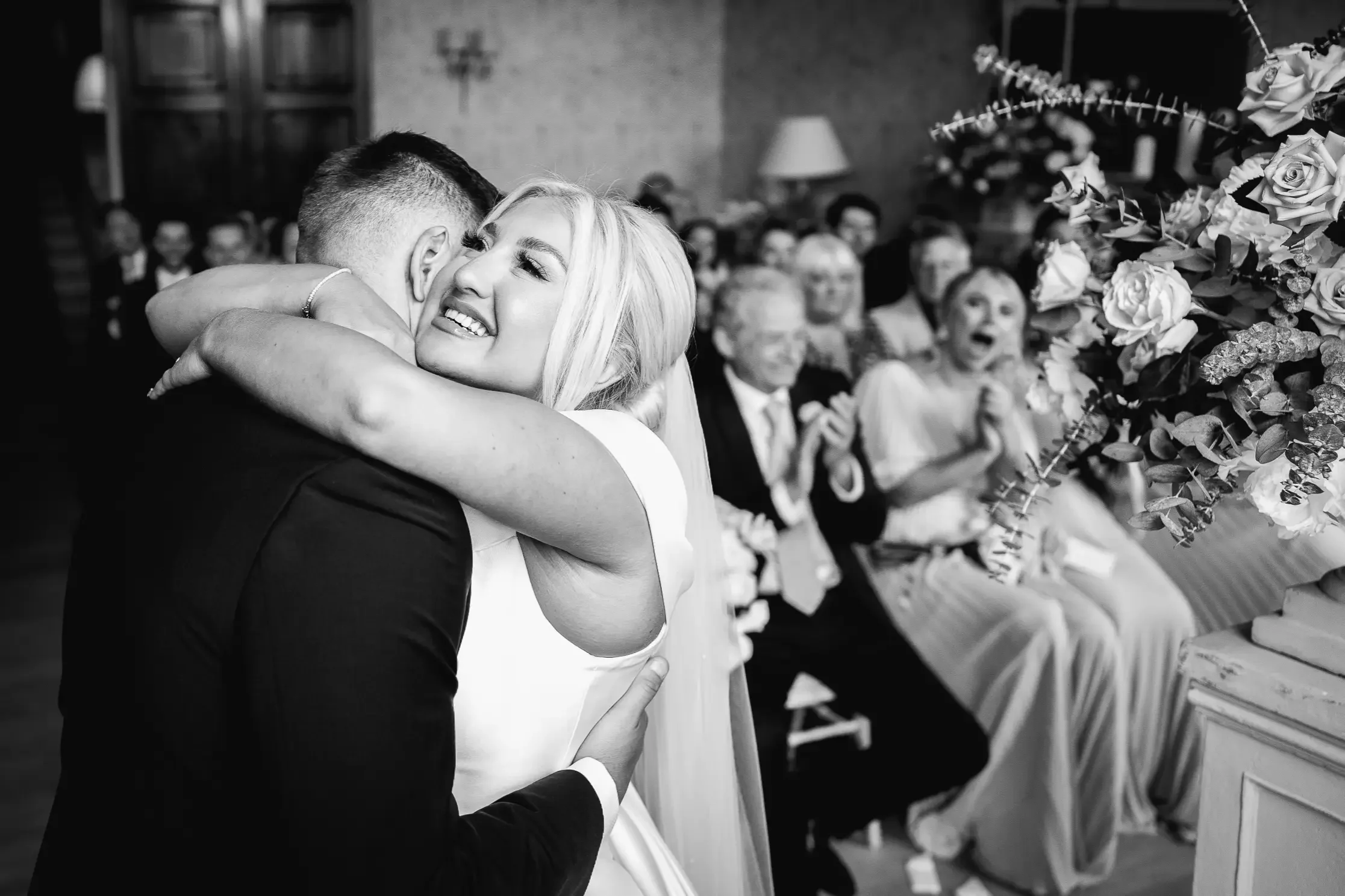 Black and white photo of a bride and groom embracing at their wedding ceremony, with guests clapping and smiling in the background.