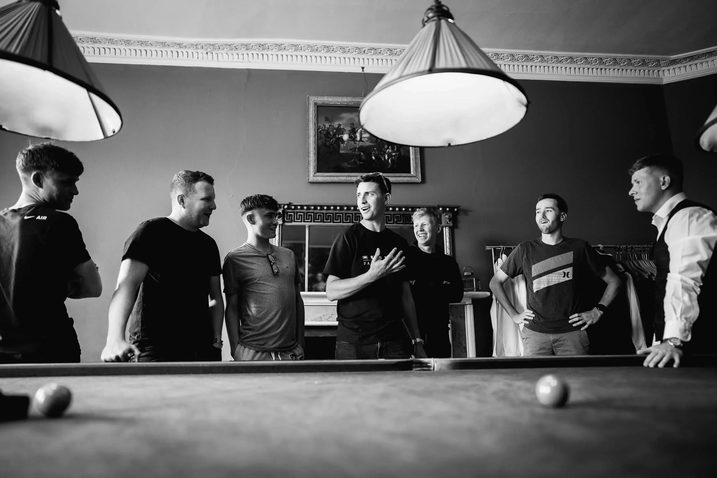 Six young men and one older man standing around a pool table in a room with ornate molding and a framed painting on the wall, engaged in conversation and laughter.