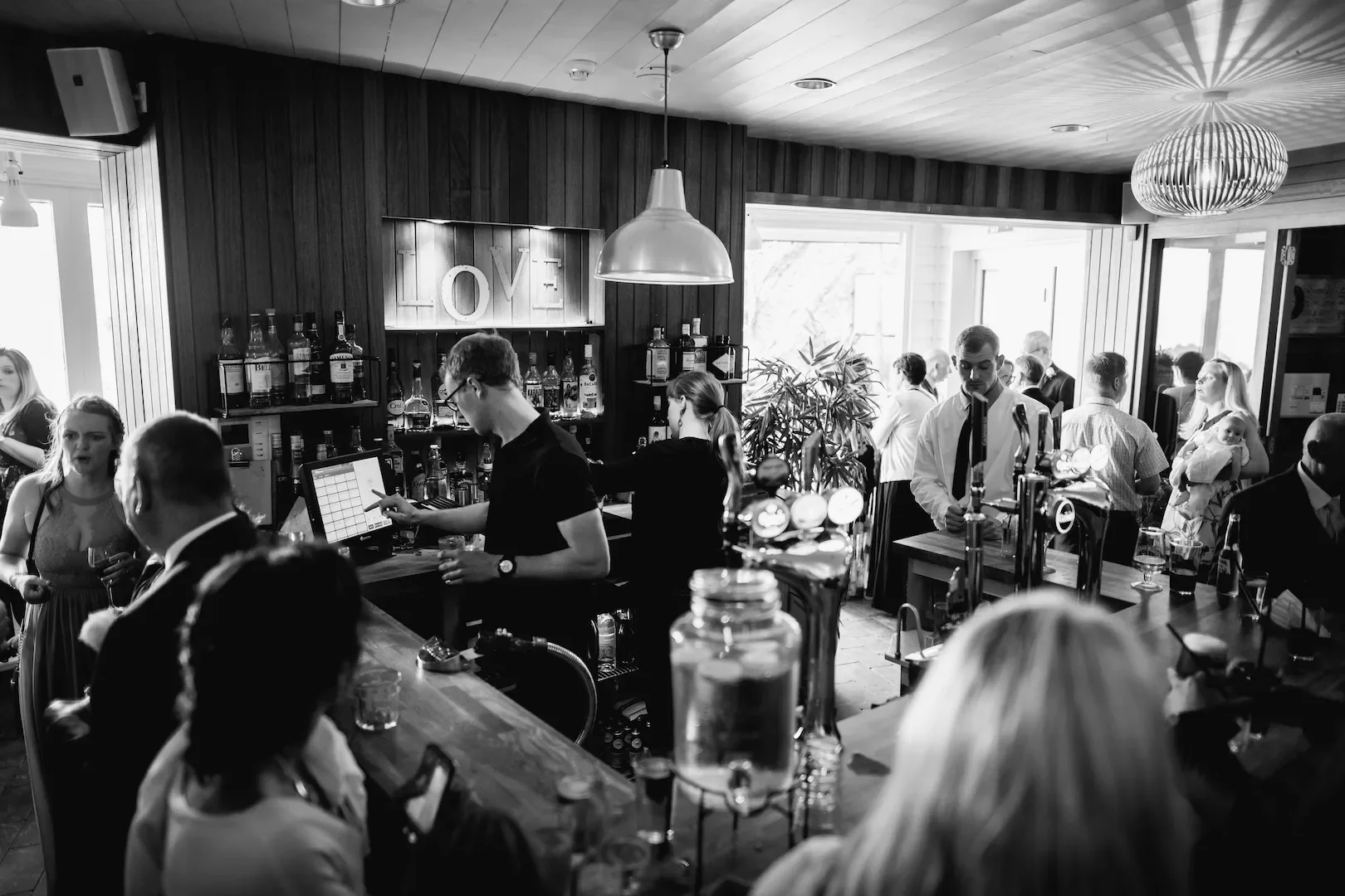 A busy bar scene with bartenders serving drinks, patrons socializing, and a large illuminated 'LOVE' sign on the dark wood-paneled wall. The room has bright natural light coming from large windows and modern decor.