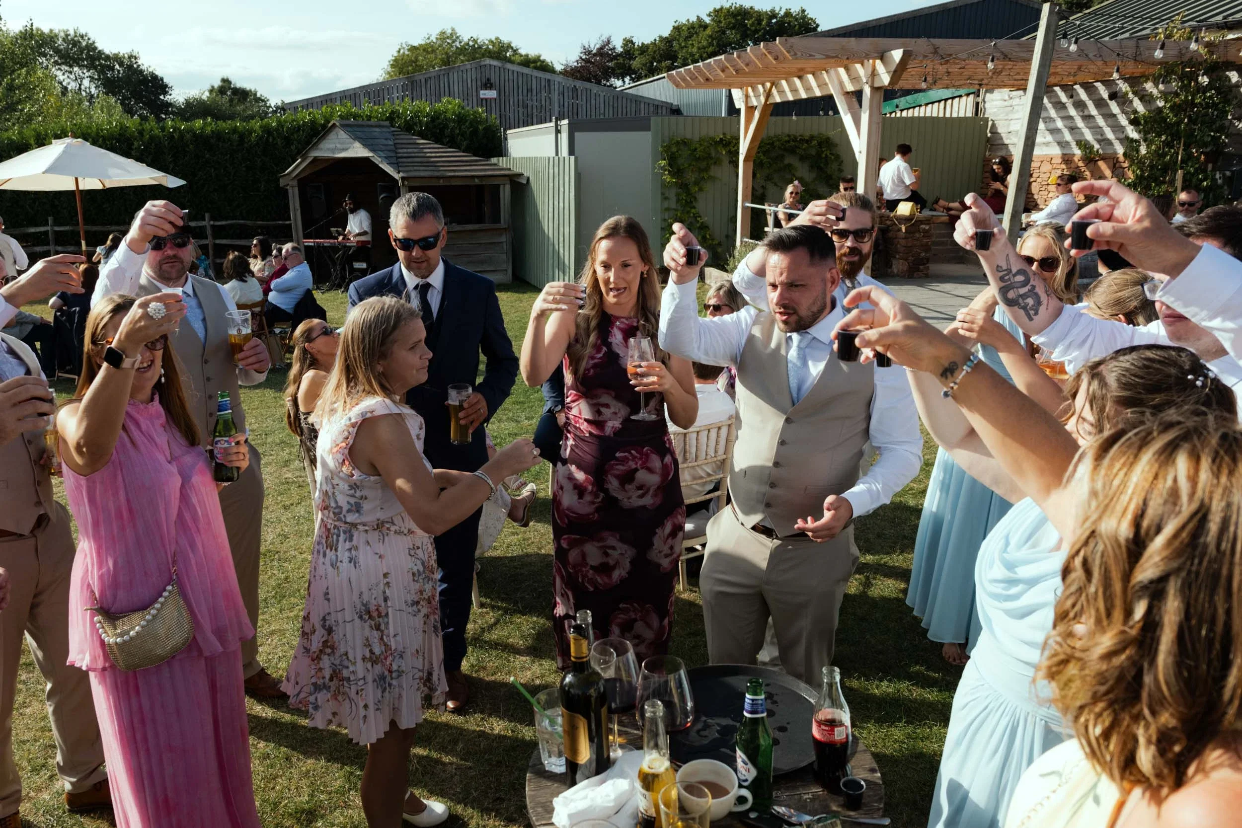 Group of people celebrating outdoors, raising glasses, with drinks on a table, at a sunny event.