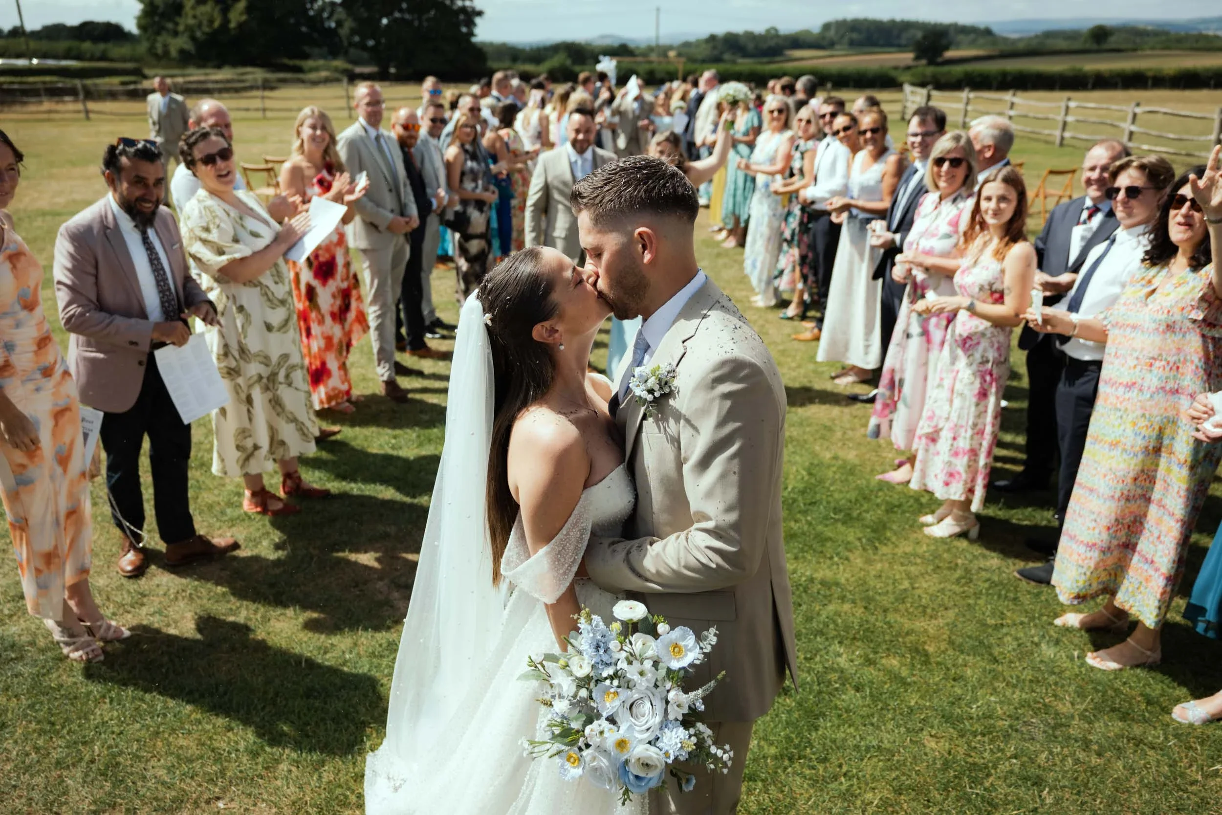 A bride and groom kiss during their outdoor wedding ceremony, surrounded by seated and standing guests in a grassy field under a clear sky.