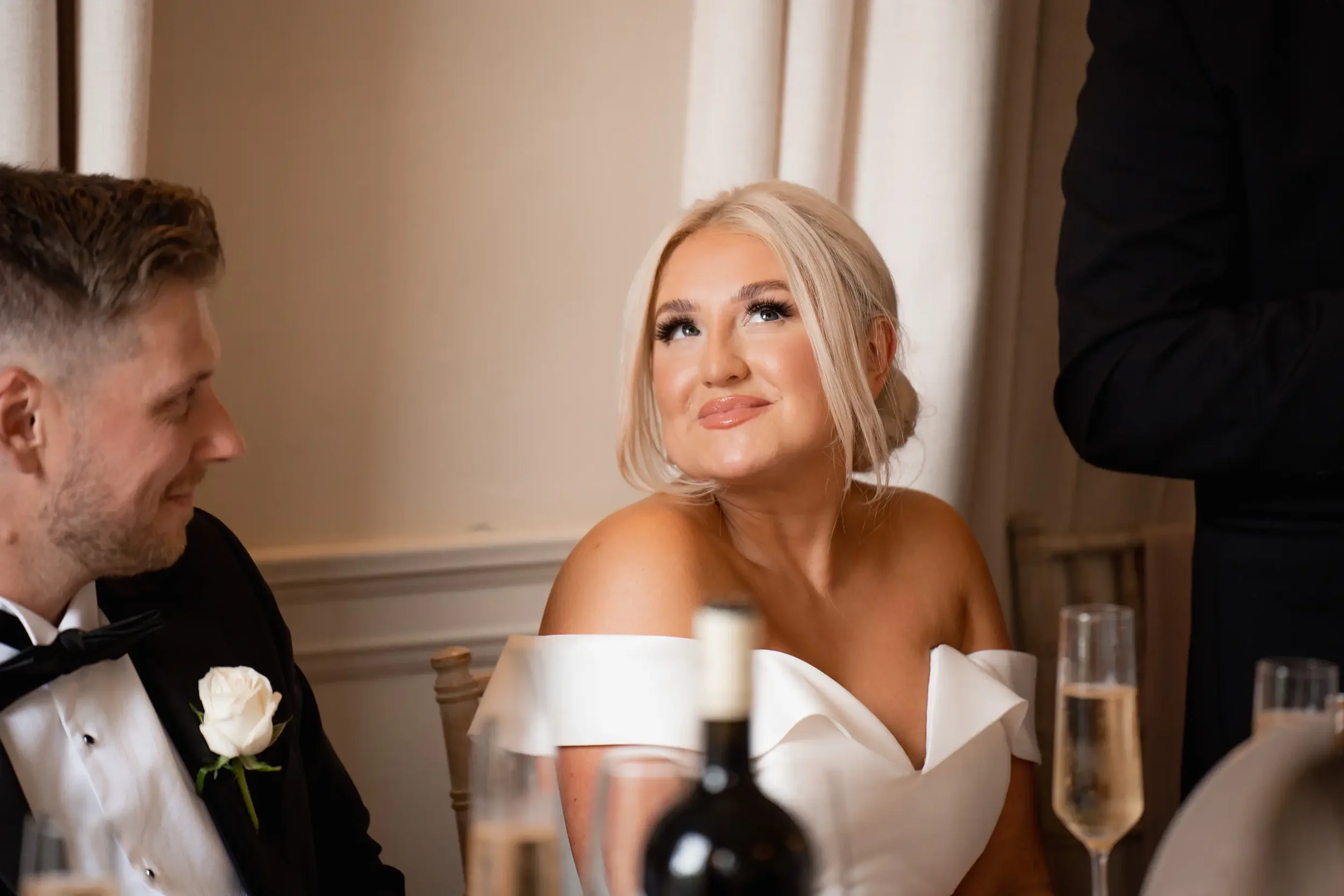 A woman with blonde hair and makeup wearing a white off-shoulder dress, sitting at a table with a man in a tuxedo, during a formal dinner event.