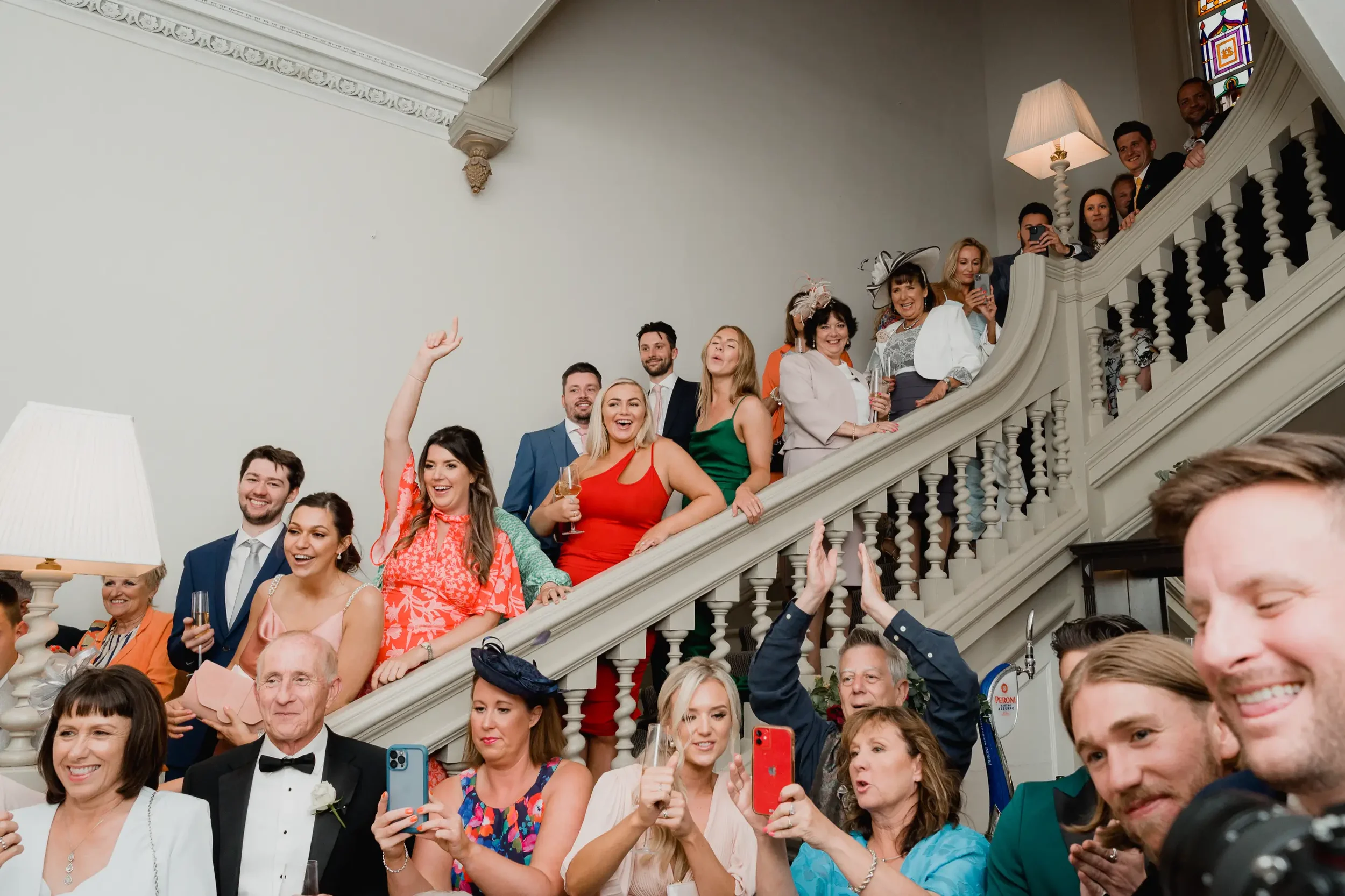 People gathered at a celebration, some on a staircase and others in front, smiling and taking photos. They are dressed in formal attire, with some holding glasses of champagne.