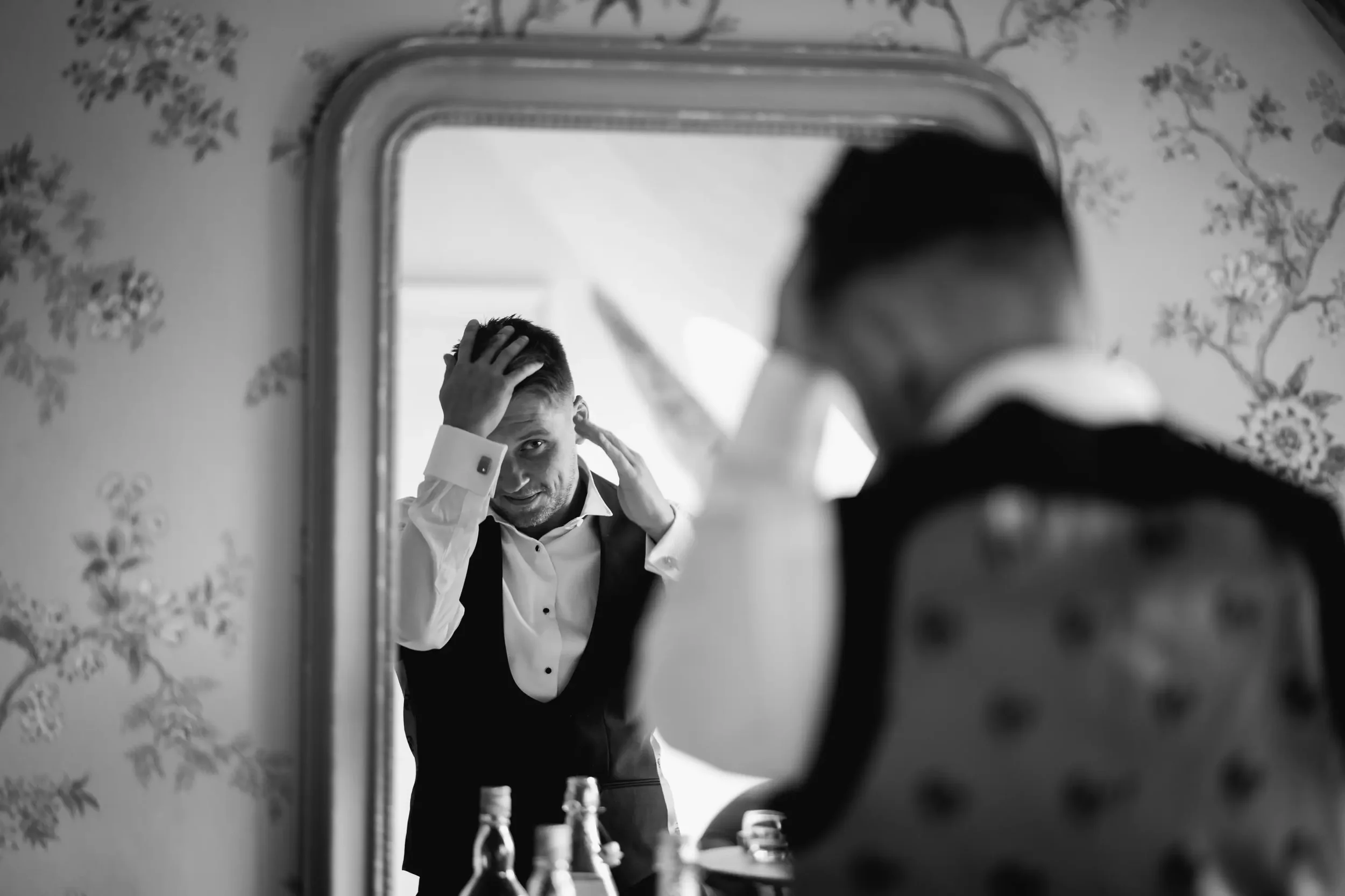 A man looking at himself in a mirror, fixing his hair. He is wearing a formal tuxedo with a white shirt.
