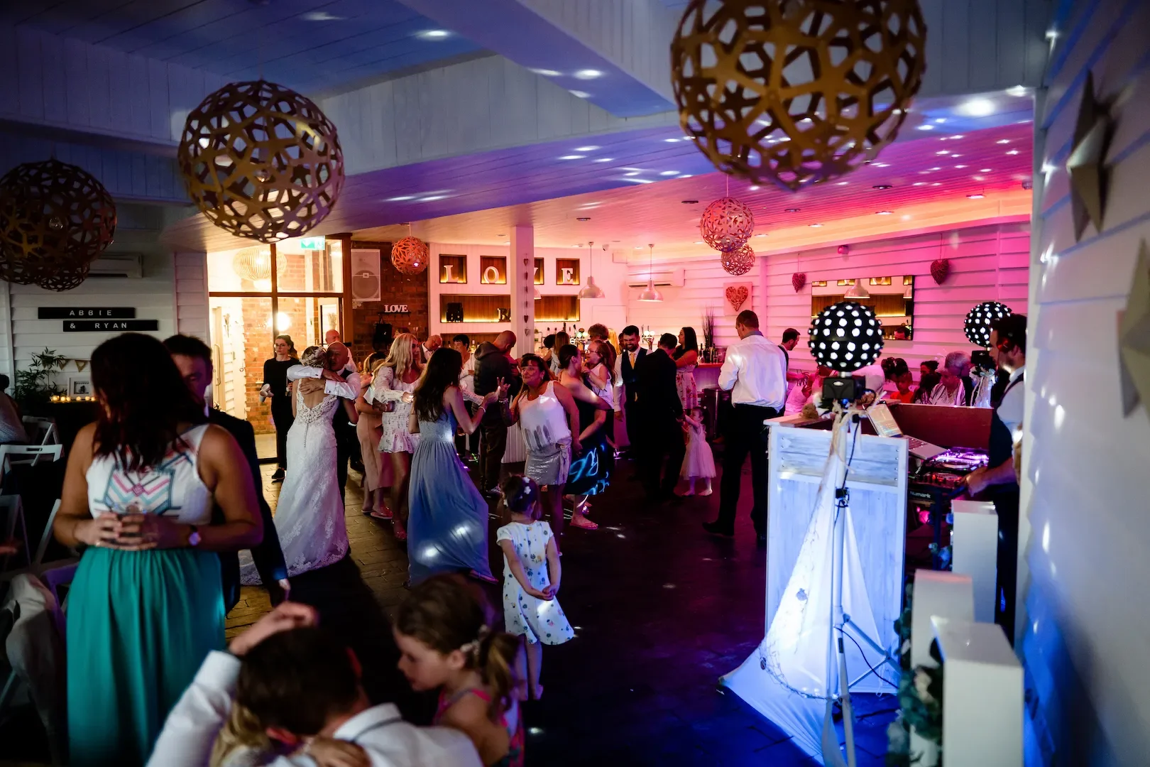 People dancing and socializing at a wedding reception inside a decorated venue with pink lighting, hanging spherical ornaments, a DJ booth, and ambient lighting.