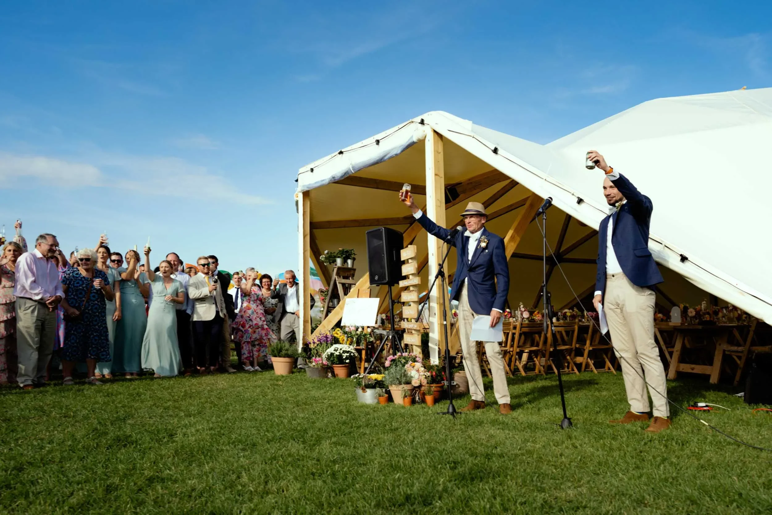 A wedding celebration outdoors with guests raising glasses, two men in suits and hats making a toast at a decorated reception tent under a blue sky.
