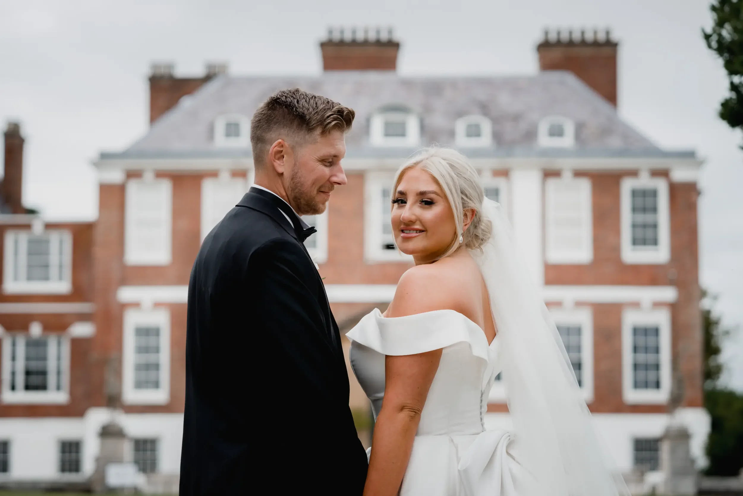Bride and groom standing outdoors in front of a large brick house, wedding attire, romantic pose.