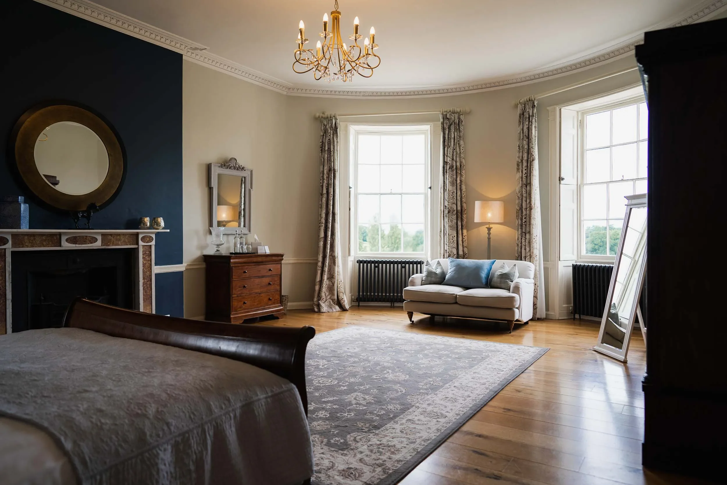 Elegant living room with large windows, wooden floors, a white couch with pillows, a side table with lamp, and a mirror on the wall.