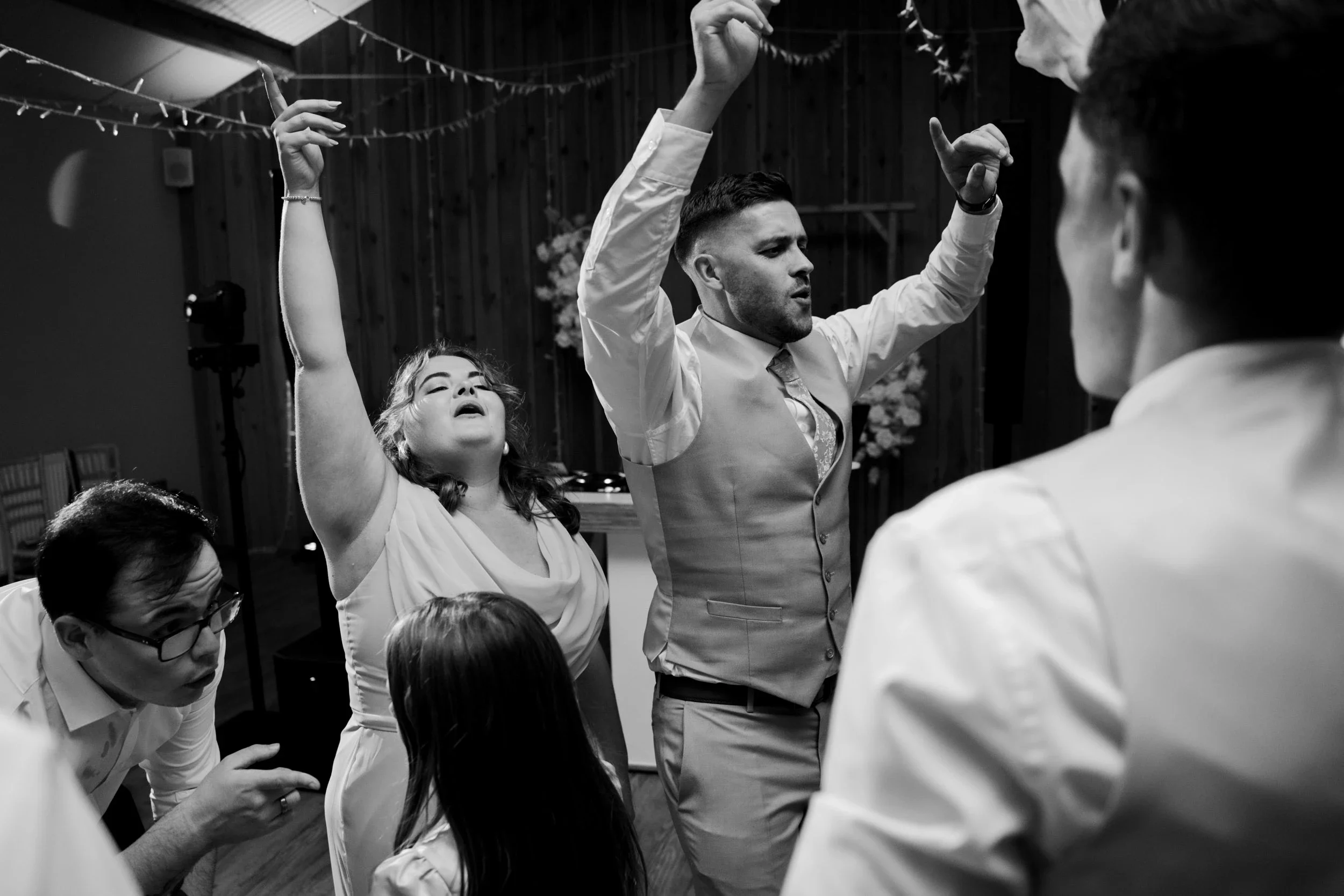 People dancing and enjoying themselves at a celebration or party, with a woman raising her arms and a man with hands in the air, in a warmly decorated indoor setting.