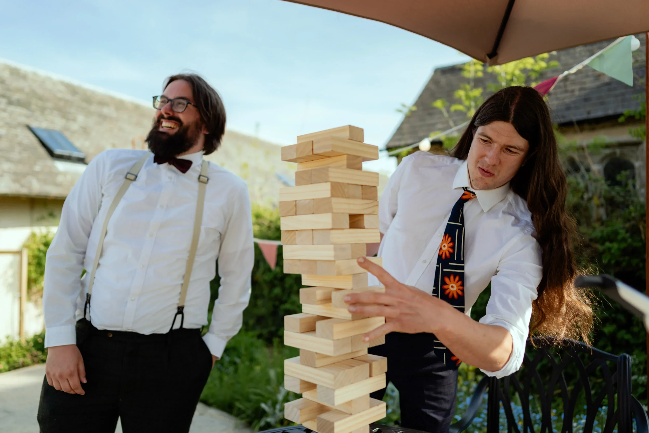 Two men in white shirts and ties playing large outdoor Jenga, one is smiling and the other is removing a wooden block, in a garden under a canopy with decorative flags.