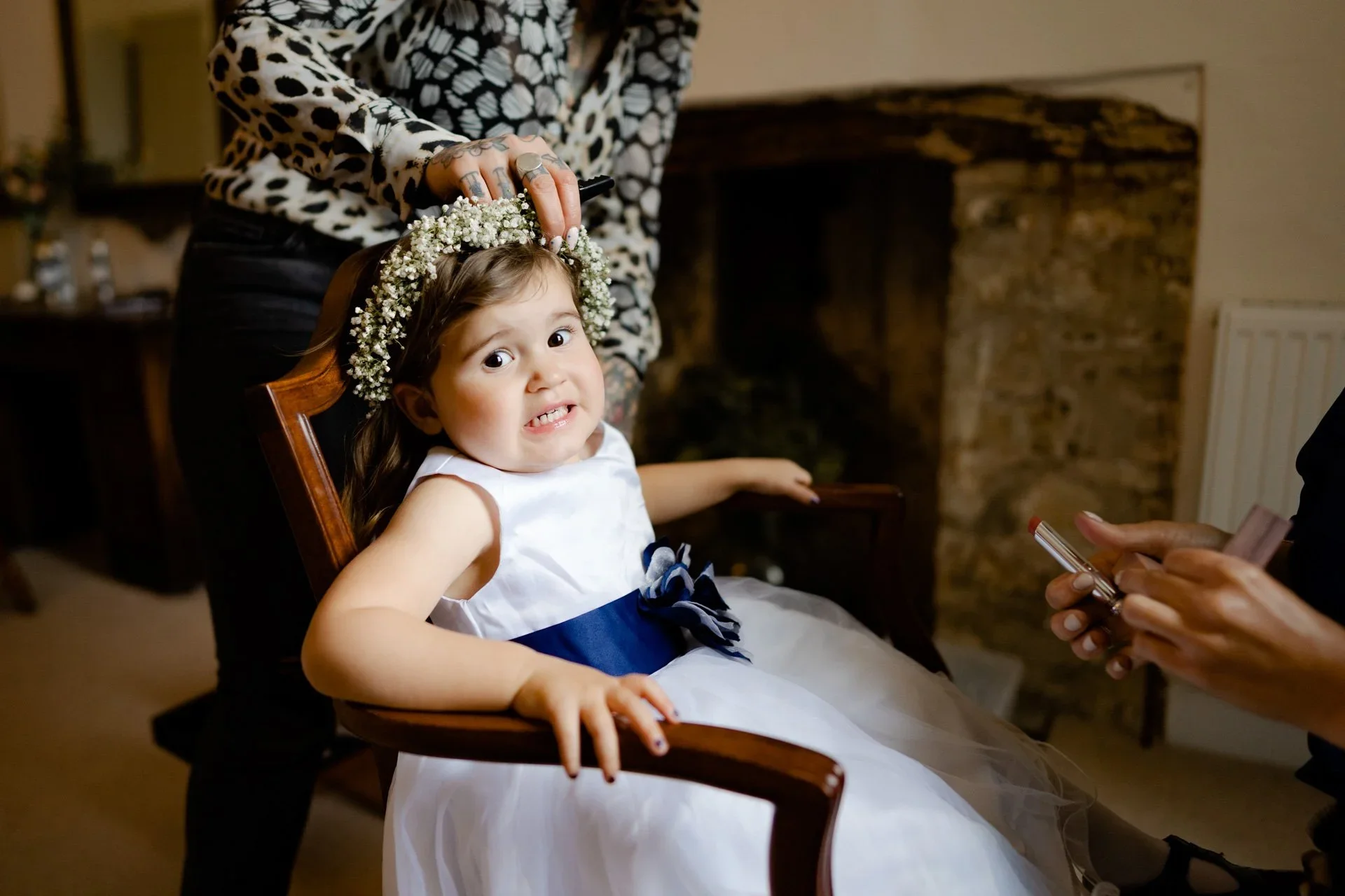 A young girl in a white dress with a navy sash sits in a wooden chair, looking nervously at the camera. A woman stands behind her, placing a floral crown on her head. Another person to the right holds a phone or camera. The setting is indoors with a stone fireplace in the background.
