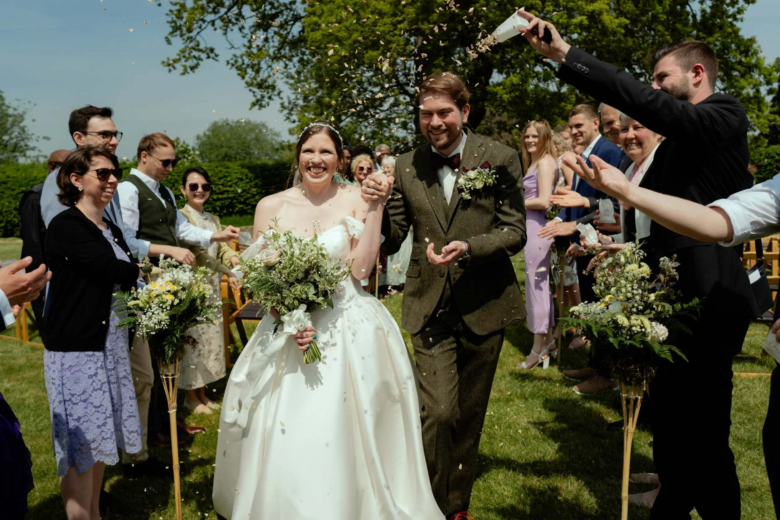 Bride and groom walking arm in arm during outdoor wedding celebration, surrounded by cheerful guests throwing flower petals.