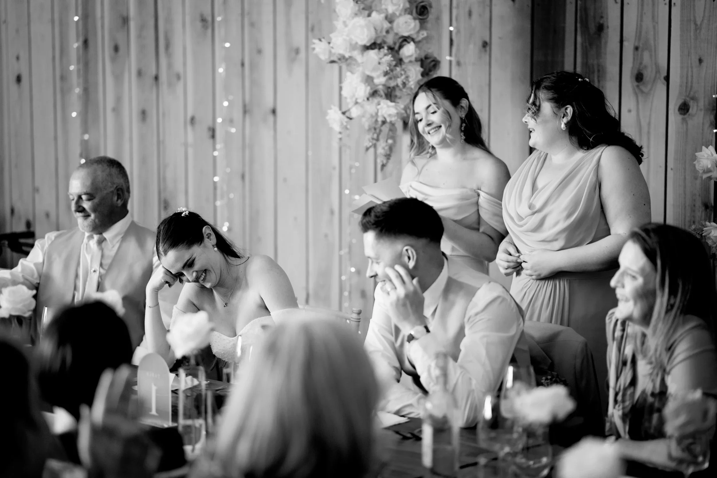 People are sitting and standing at a wedding reception, smiling and laughing. A woman with dark hair and a strapless dress is in the foreground, laughing with her hand on her head. Behind her, a man with short dark hair is sitting, smiling and touchi