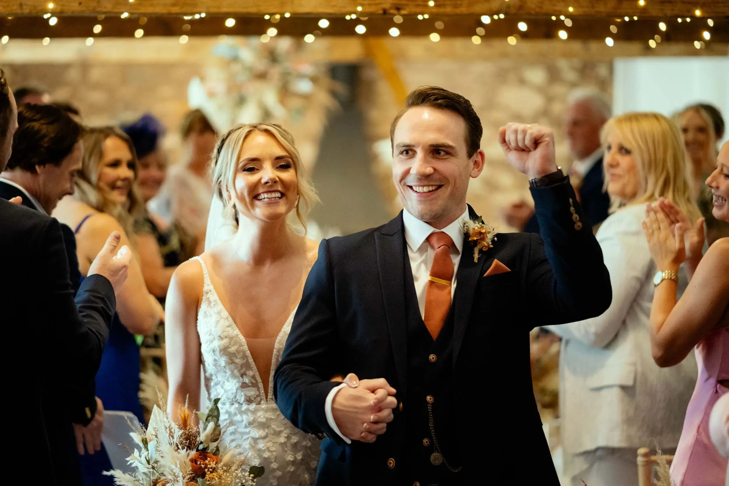 Happy bride and groom at wedding celebration, surrounded by guests, with rustic interior decorated with string lights.
