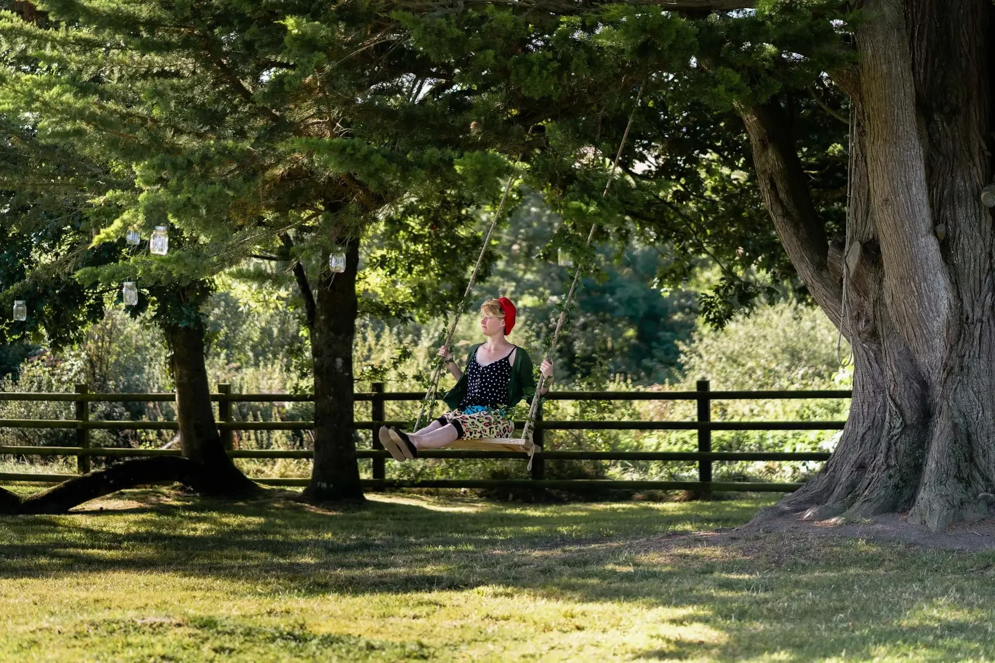 A woman with blonde hair, wearing a red headscarf, a black and white polka dot top, leopard print pants, and a black cardigan, sitting on a wooden swing hanging from a large tree in a park.