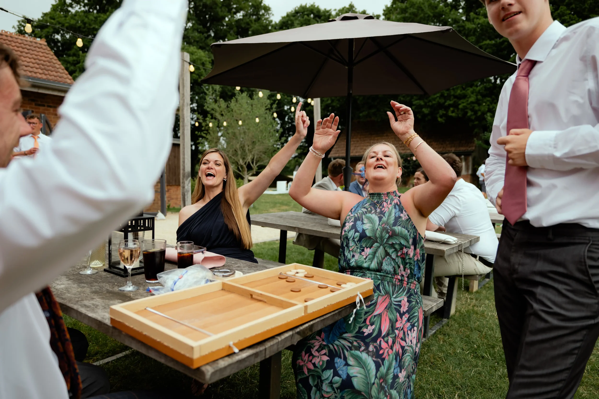 People celebrating and playing a game outdoors at a social gathering, with drinks on the table and a large umbrella overhead.