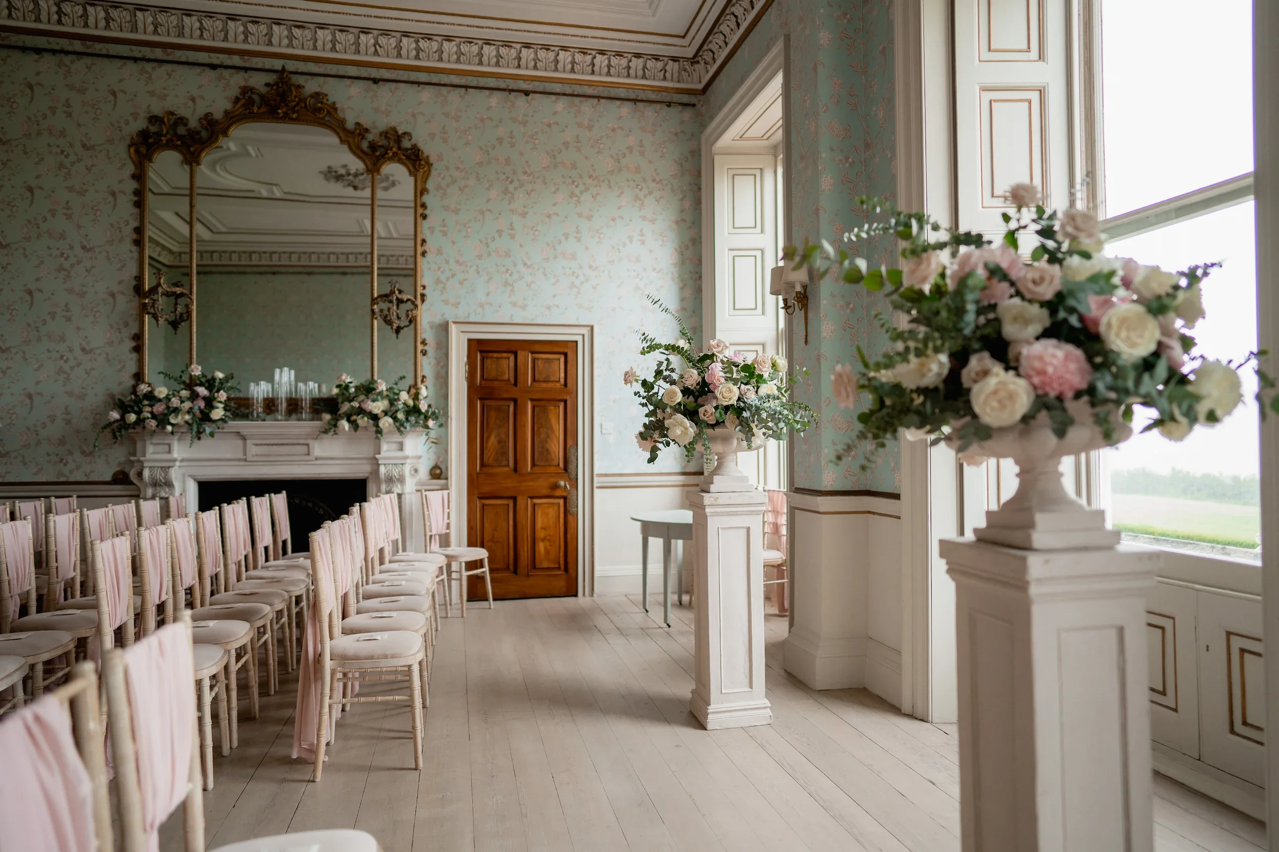 Elegant room with floral arrangements on pedestals, rows of chairs with pink ribbons, and a large mirror above the fireplace.