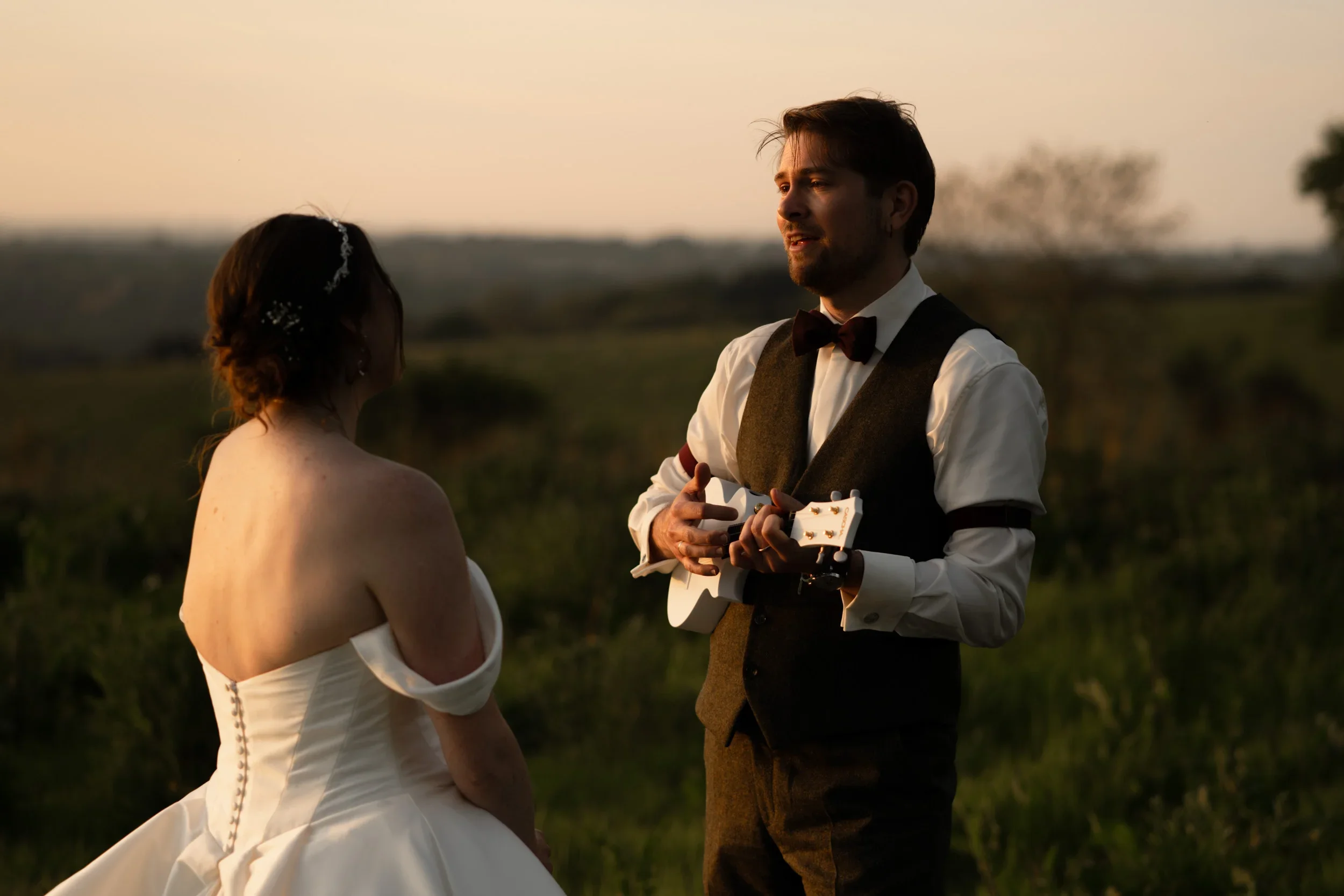 A couple on their wedding day standing outdoors during sunset. The bride is facing the groom, wearing a white off-shoulder wedding dress with buttons down the back. The groom is holding a small white guitar, dressed in a white shirt, dark vest, dark 