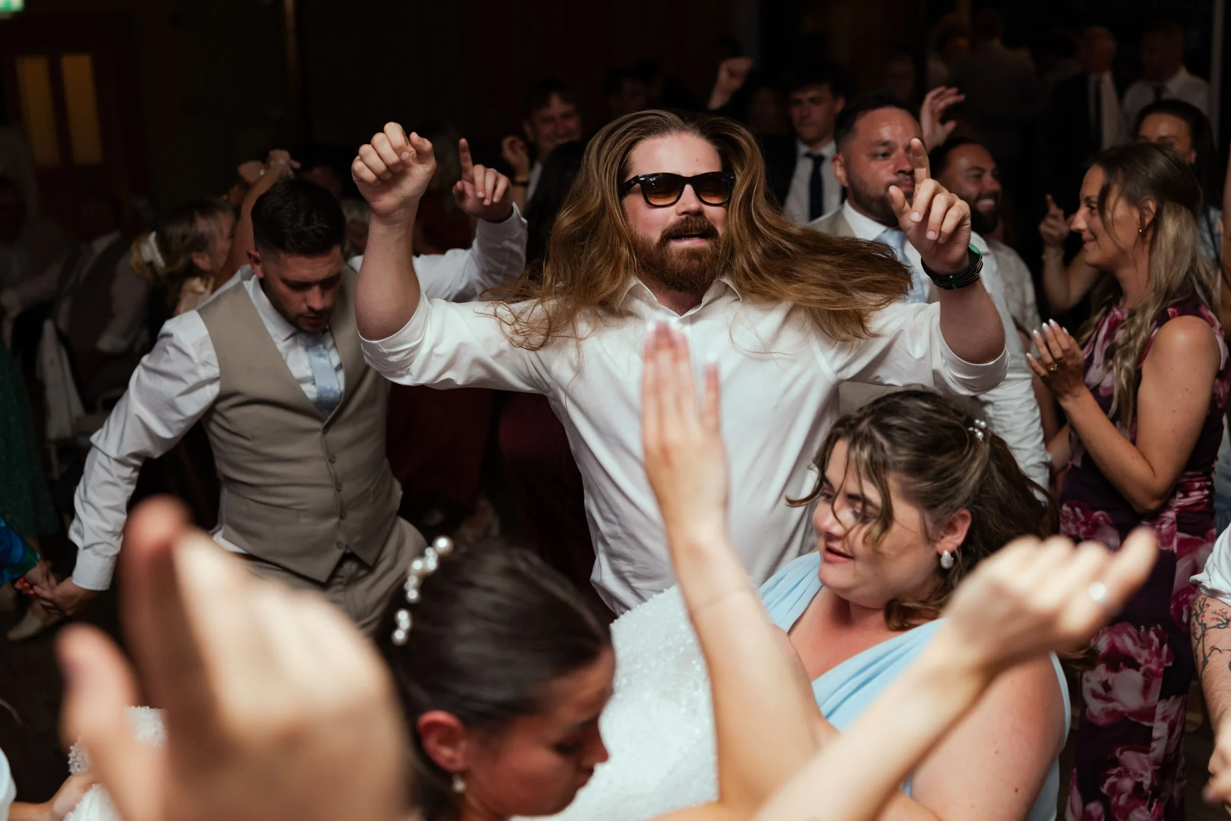 People dancing and celebrating at a party or wedding reception. A man with long hair, beard, sunglasses, and a white shirt is dancing with his arms raised. Several other people are smiling, clapping, and enjoying the event.