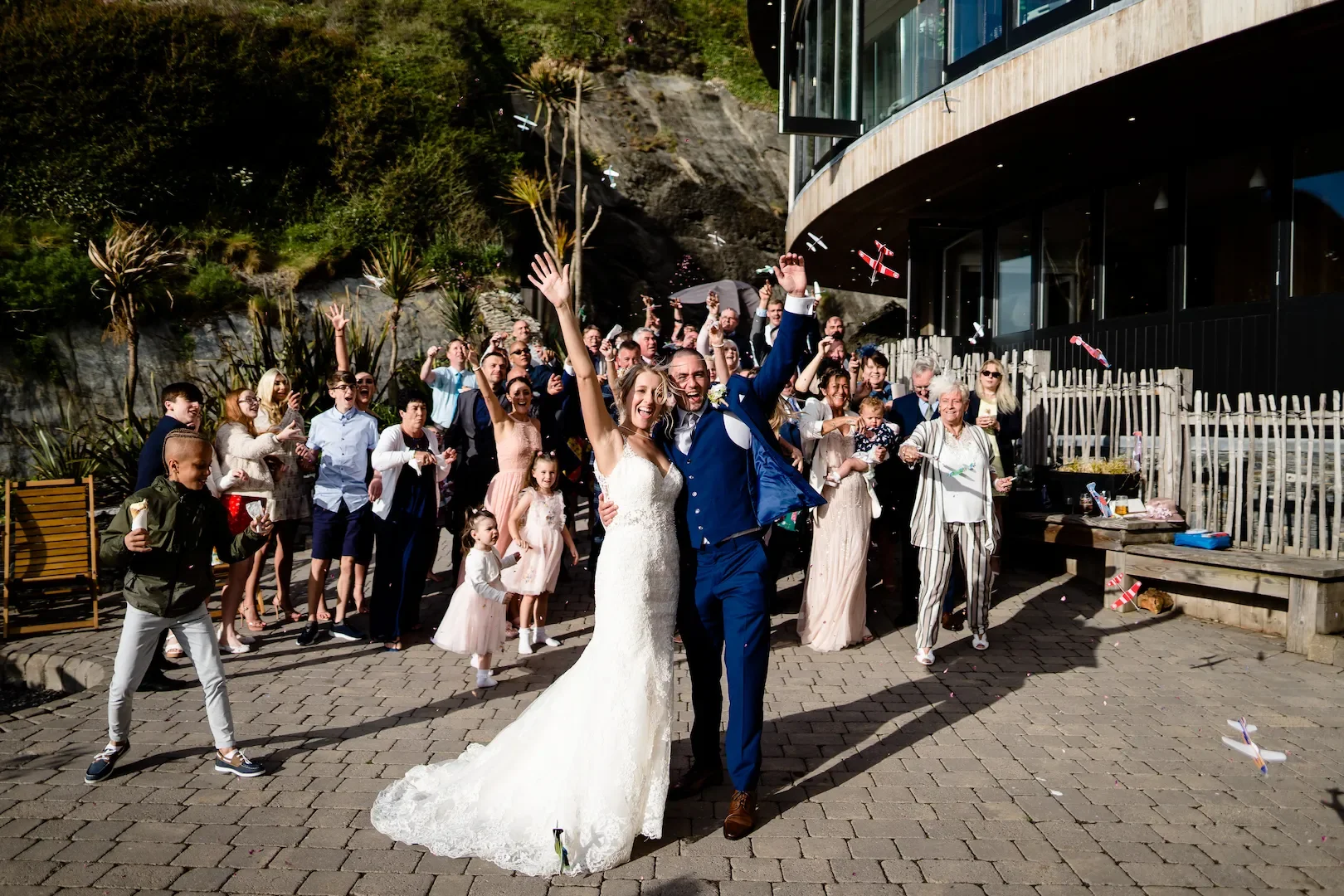 A wedding celebration outdoors with a diverse group of guests cheering and throwing confetti, including a smiling bride in a white gown and groom in a blue suit.