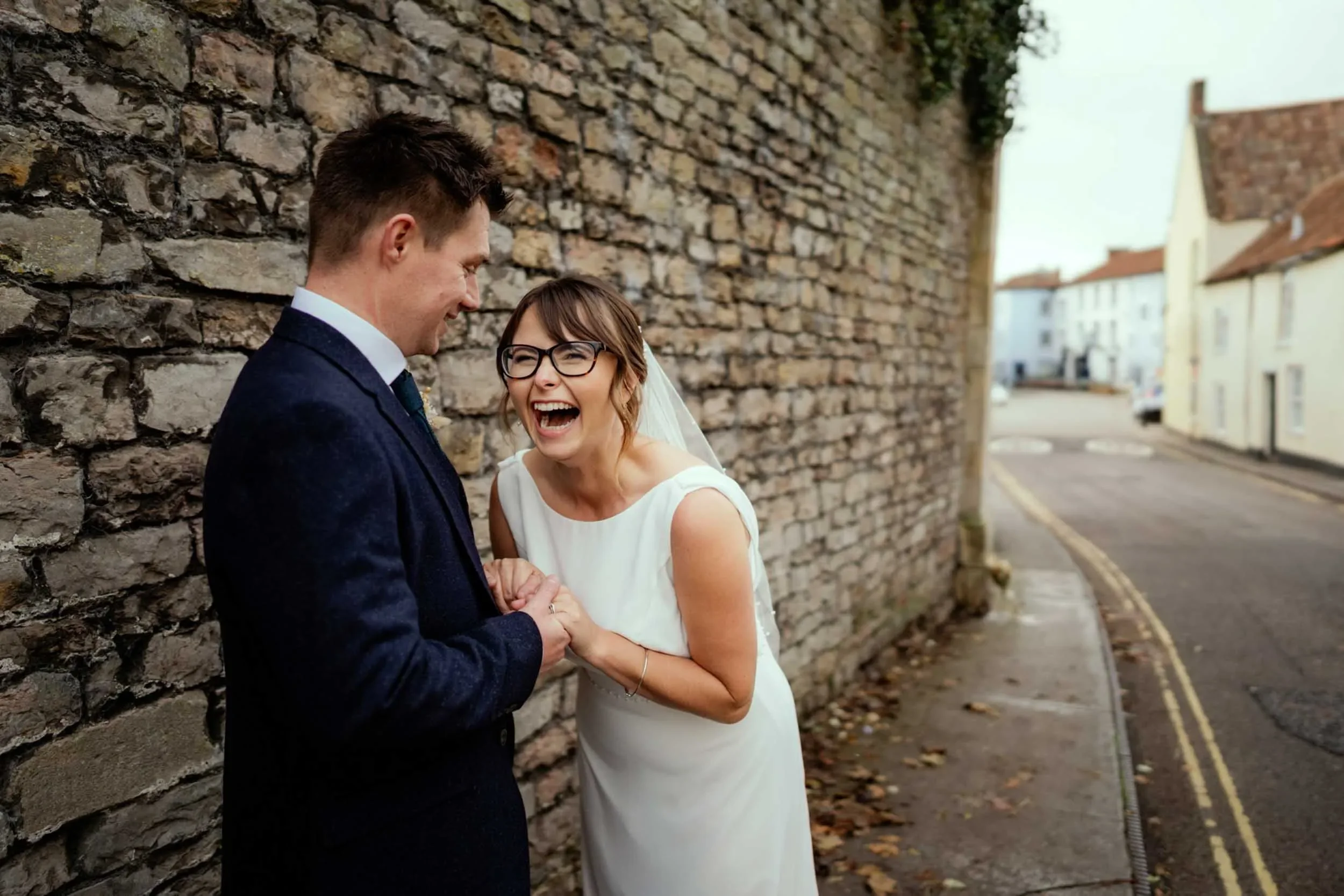 A smiling bride in a white dress and glasses holding hands with a groom in a dark suit, standing against a brick wall on a street with houses in the background.