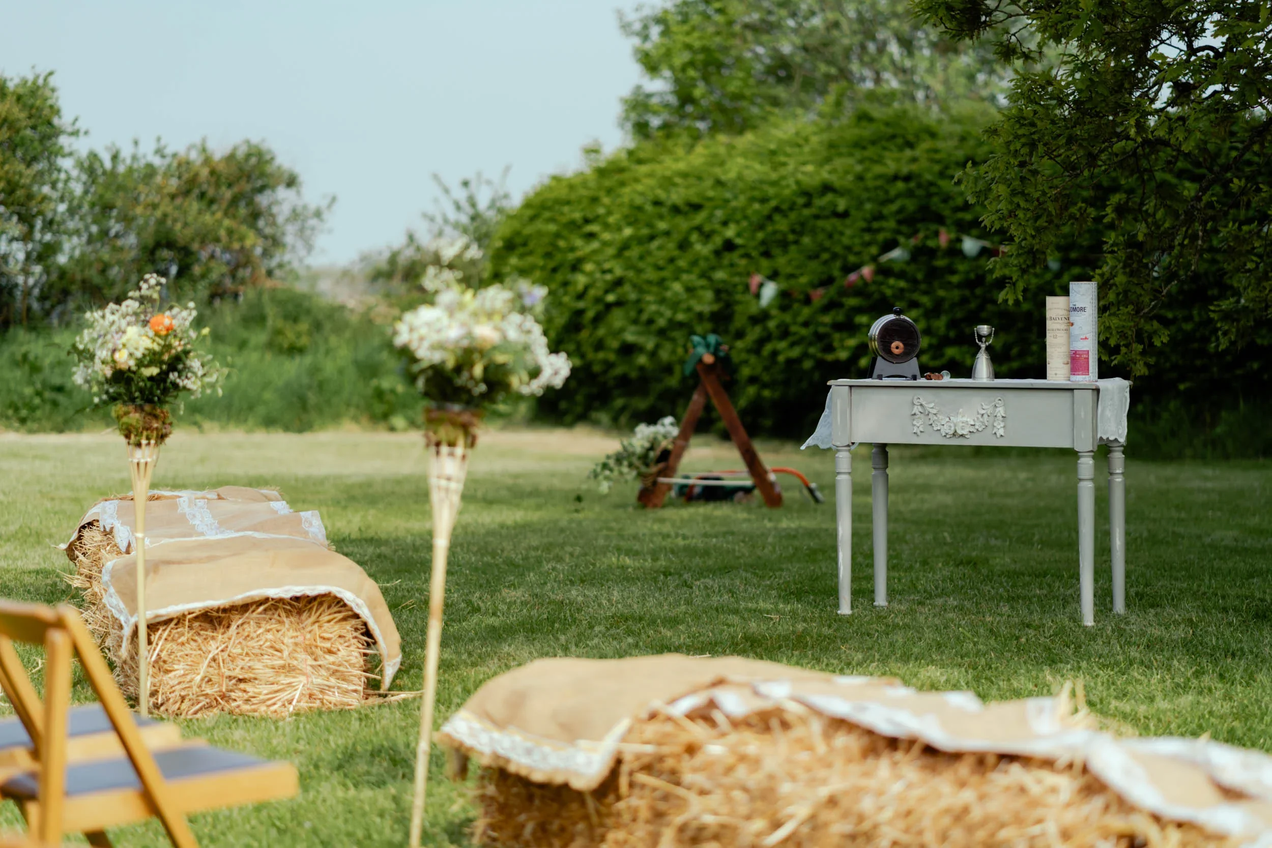 Outdoor garden scene with hay bales covered with cloth, flower arrangements, a white decorated table with items on top, and a green hedge in the background.