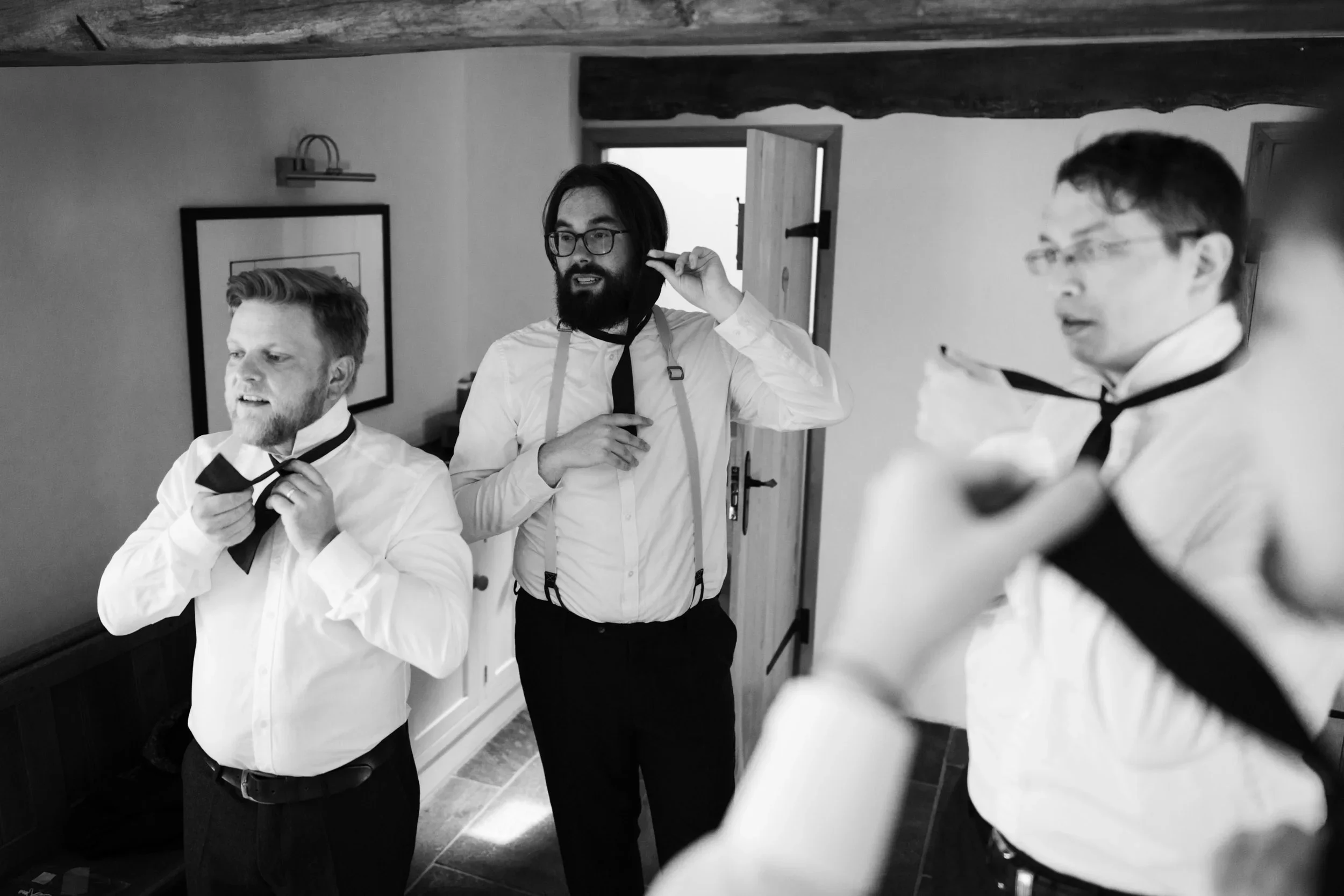 Three men in white shirts and black ties getting ready for a formal event, adjusting their ties, indoors with a wooden door and picture on the wall.